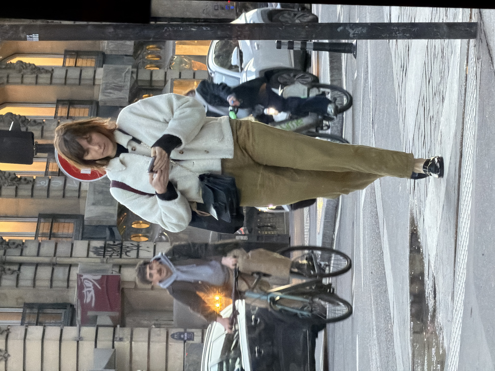 The image depicts an urban street scene in Paris, France, during what appears to be dusk or early evening conditions.
In the foreground, a woman with medium-length brown hair is walking towards the viewer's right, positioned on a grey paved sidewalk. She is wearing a cream-colored sherpa jacket over a black long-sleeved top, olive-green tailored trousers, and black patent leather loafers with gold hardware. A black shoulder bag with a gold chain strap hangs across her body. She is actively engaged with a silver smartphone held in both hands, her gaze directed downwards at the screen.
Behind her, on the street, multiple individuals and vehicles are in motion. To the left, a male with a dark mustache, wearing a grey hooded sweatshirt under a dark jacket and tan trousers, is riding a light blue road bicycle. The bicycle's front light is illuminated, creating a bright flare. Behind this cyclist, a dark grey sedan or SUV is partially visible, moving in the same direction.
Further to the right, a second cyclist, dressed in dark attire, is riding a bicycle with a visible green accessory on its handlebars. This individual is partially obscured by the main woman and a black street pole on the far right of the frame. A white SUV is visible in the background, behind this second cyclist.
The environment includes classic Parisian Haussmannian-style buildings with light-colored stone facades, ornate details, and multi-paned windows, some of which are illuminated with warm orange light. Graffiti is present on some building surfaces and a red circular "no entry" sign is visible in the mid-ground. The street is paved with grey asphalt, featuring white traffic markings, and a small puddle is present on the road surface near the foreground sidewalk.