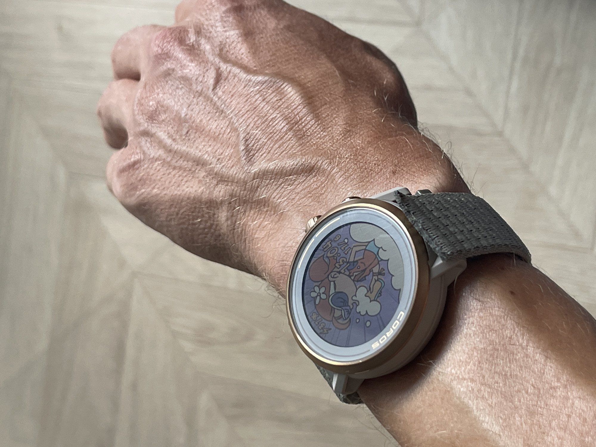 The media file shows a close-up of a person's arm wearing a smartwatch. The smartwatch has a circular face with a colorful cartoon graphic displayed on it, featuring characters with large eyes and smiling faces. The watch band is a dark gray woven fabric. Visible text on the watch face indicates the time as "15:04" and "17:0". The brand name "COROS" is also visible along the bottom edge of the watch face. The person's arm is tanned and shows prominent veins and fine hairs. The background appears to be a light-colored, textured surface, possibly wood flooring, with diagonal lines suggesting planks. The lighting suggests it is daytime.