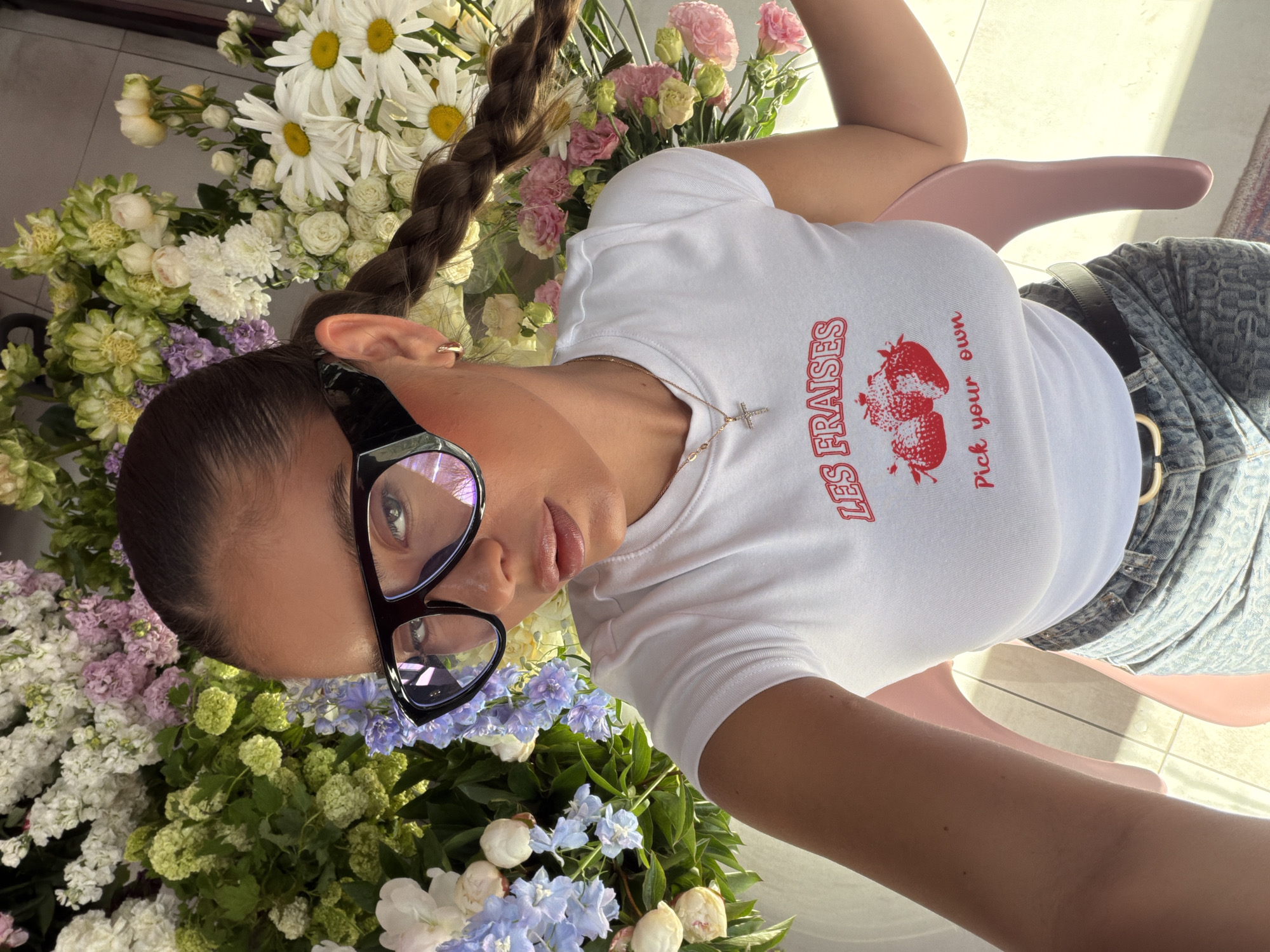 A woman wearing a white t-shirt with a red strawberry print, jeans, and a black belt, along with black glasses and a gold necklace with a cross, is surrounded by a large bouquet of flowers. She is sitting on a pink chair. Her hair is styled in a braid. The location appears to be indoors with tiled flooring. There is no indication of the city of Krasnoyarsk, Russia, in the provided image.
