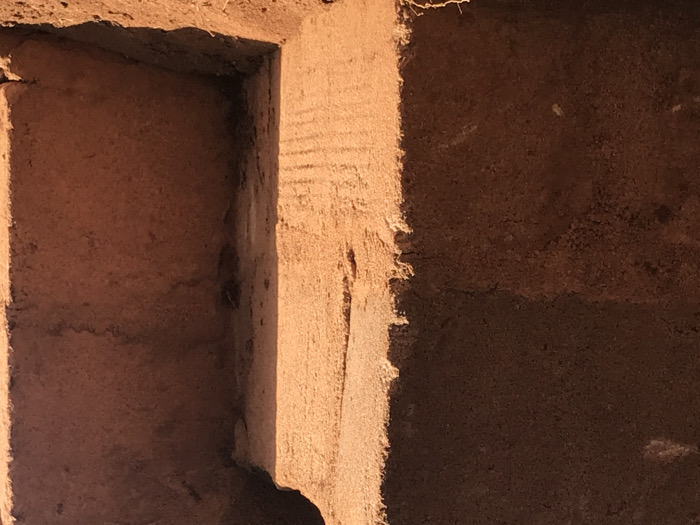 A rough, earthy structure features a square recess or niche in the left. The niche's interior and surrounding walls are textured, with varying tones of brown and tan. Light casts a strong shadow on one side, highlighting the uneven surface. Faint horizontal lines are visible on the wall beside the niche. The overall impression is of an aged, hand-built structure.