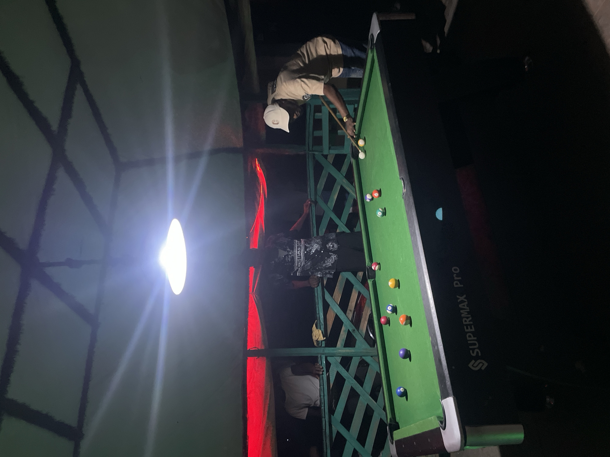 Two people play pool on an elevated outdoor green pool table at night in Lokoja, Nigeria. One person, wearing a white cap, khaki shirt, and jeans, stands over the table, holding a pool stick and aiming at a white ball. Another person, wearing a black shirt and pants, is below, next to the table. The "SUPERMAX PRO" brand is printed on the side of the pool table. Red lighting accents the edge of a building or structure. The location is outdoors, underneath a lattice-like structure. A light above casts a glare on the scene.