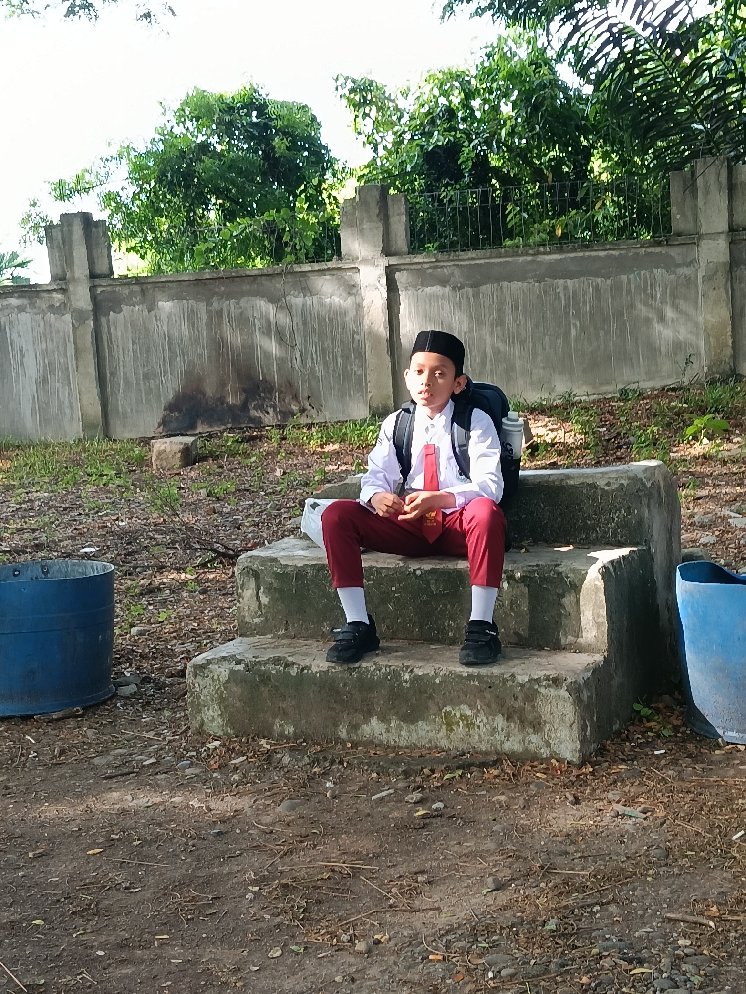 The image shows a young boy in a school uniform sitting on concrete steps in an outdoor environment. The boy wears a white shirt, red tie and pants, white socks, black shoes, and a black peci hat. He also has a backpack on. He is sitting with his hands clasped in his lap. The background includes a weathered concrete wall, trees, and two blue plastic barrels. The ground is littered with leaves and small debris. The location is Lhokseumawe, Indonesia.
