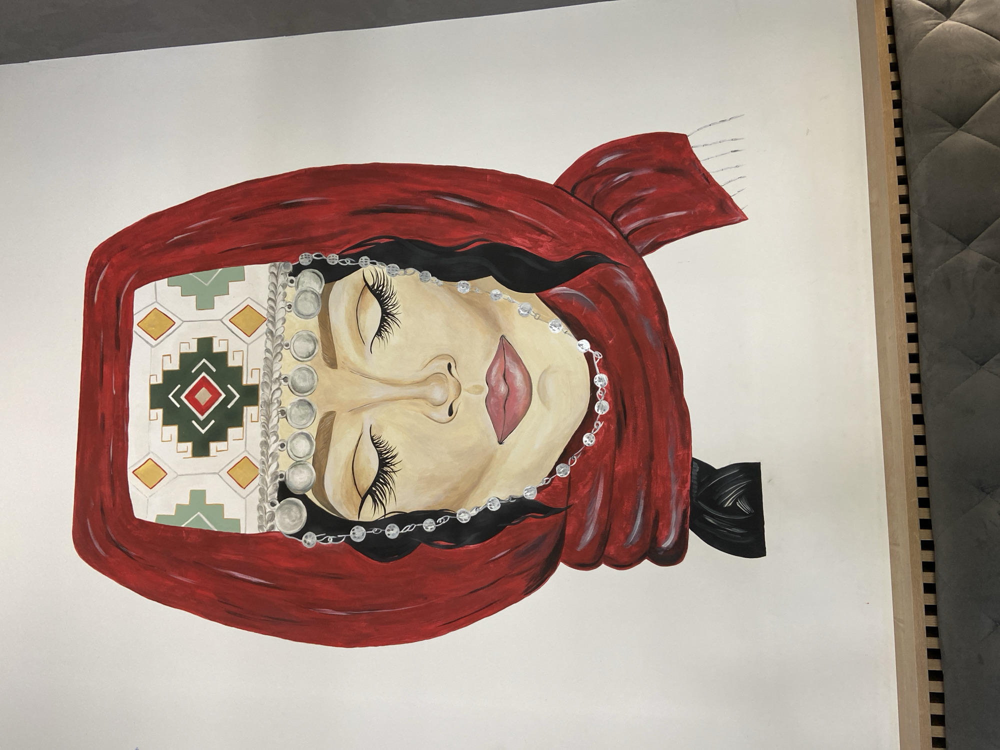 A painting of a woman wearing a red headscarf and traditional headdress, with eyes closed and adorned with silver jewelry, is displayed against a white background. The woman's face is rendered in shades of tan and pink, with dramatic eyelashes and full lips. Her headdress features intricate geometric patterns in white, green, yellow, and red. Silver coins and beaded chains hang from the headdress, framing her face. A thick red scarf is wrapped around her head and neck, with its ends extending down. Her dark hair is visible in a braid beneath the scarf. The painting appears to be on a large canvas, with its lower edge resting on a piece of furniture with a patterned upholstered surface and a slatted wooden trim. The setting appears to be indoors, possibly a home or gallery space in the city of Brest, Belarus.