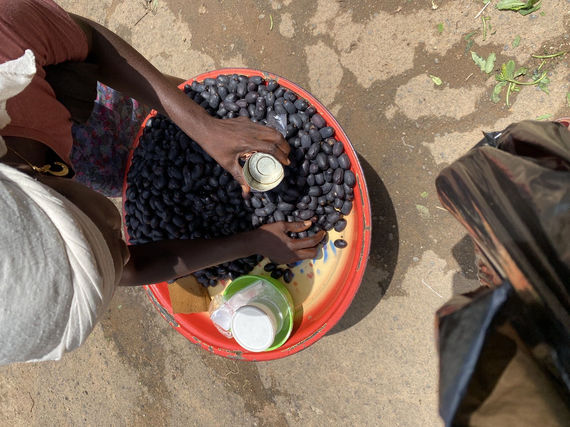 In Jos, Nigeria, an individual is tending to a large quantity of dark-colored, oval-shaped fruits, possibly olives or a similar local variety, arranged in a wide, shallow red bowl. The fruits fill most of the bowl, with some spilled onto the surrounding surface. The individual, wearing a white headscarf and a reddish shirt, is using a small metallic cup to scoop or measure the fruits. A green bowl containing two white containers, possibly jars or cups, sits within the larger red bowl. The scene appears to be outdoors, with a dusty, uneven ground surface visible around the bowl. A portion of a black plastic bag is also visible in the foreground, suggesting a market or roadside stall setting. The lighting indicates it is daytime, with shadows cast on the ground.