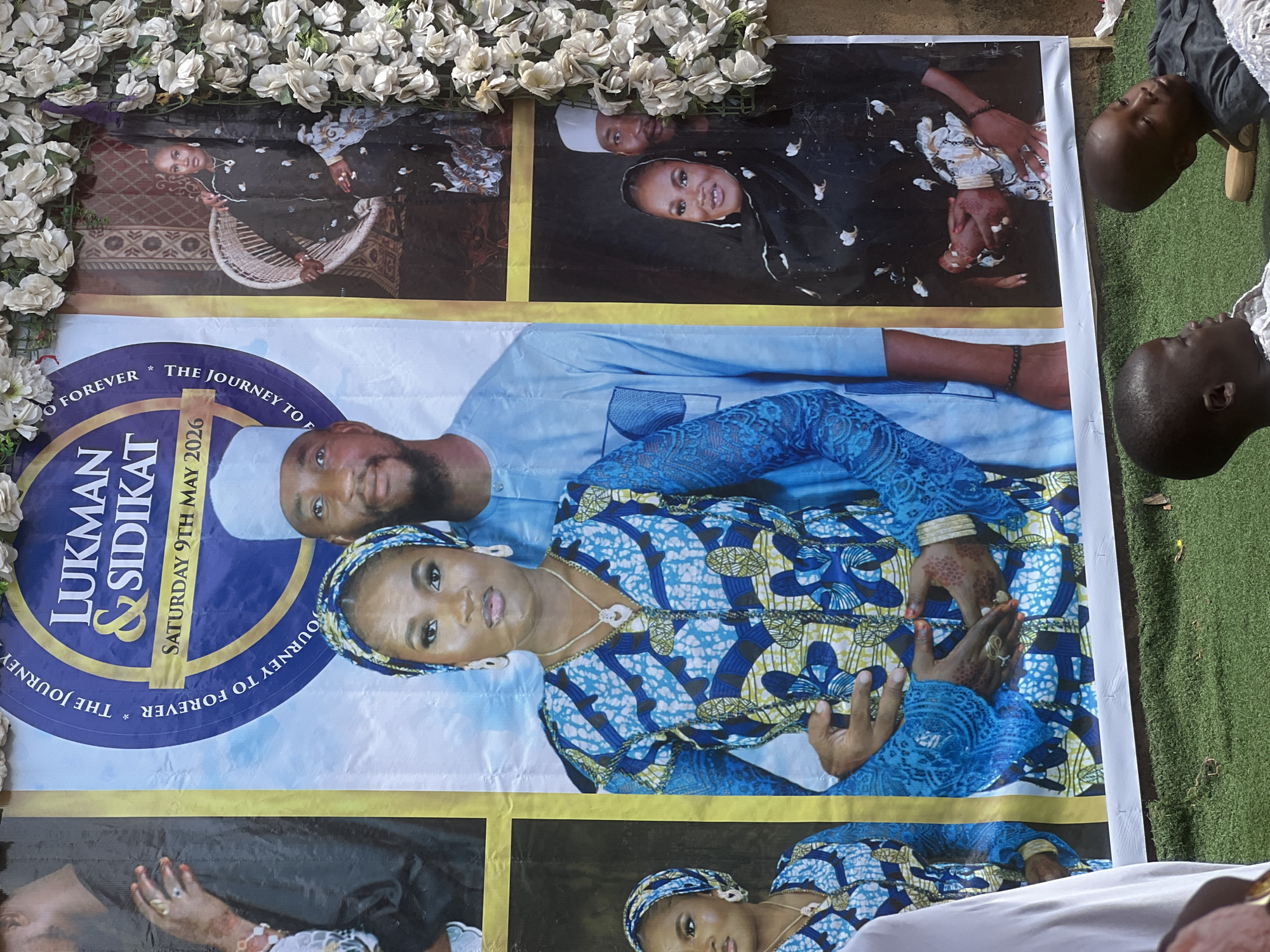 A rectangular poster, potentially laid on the ground outdoors, appears to commemorate a wedding or similar event. The poster features several images of a couple, along with the names "Lukman & Sidikat" and the date "Saturday 9th May 2026." The overall color scheme of the poster is predominantly blue and yellow. The poster appears to be bordered by a garland of artificial white flowers along the top. Portions of people are visible along the periphery of the image, suggesting the presence of others at the scene. The poster is likely located outdoors with grass visible nearby. The city and country of Rigasa, Nigeria is stated in the prompt, but not identifiable in this image.