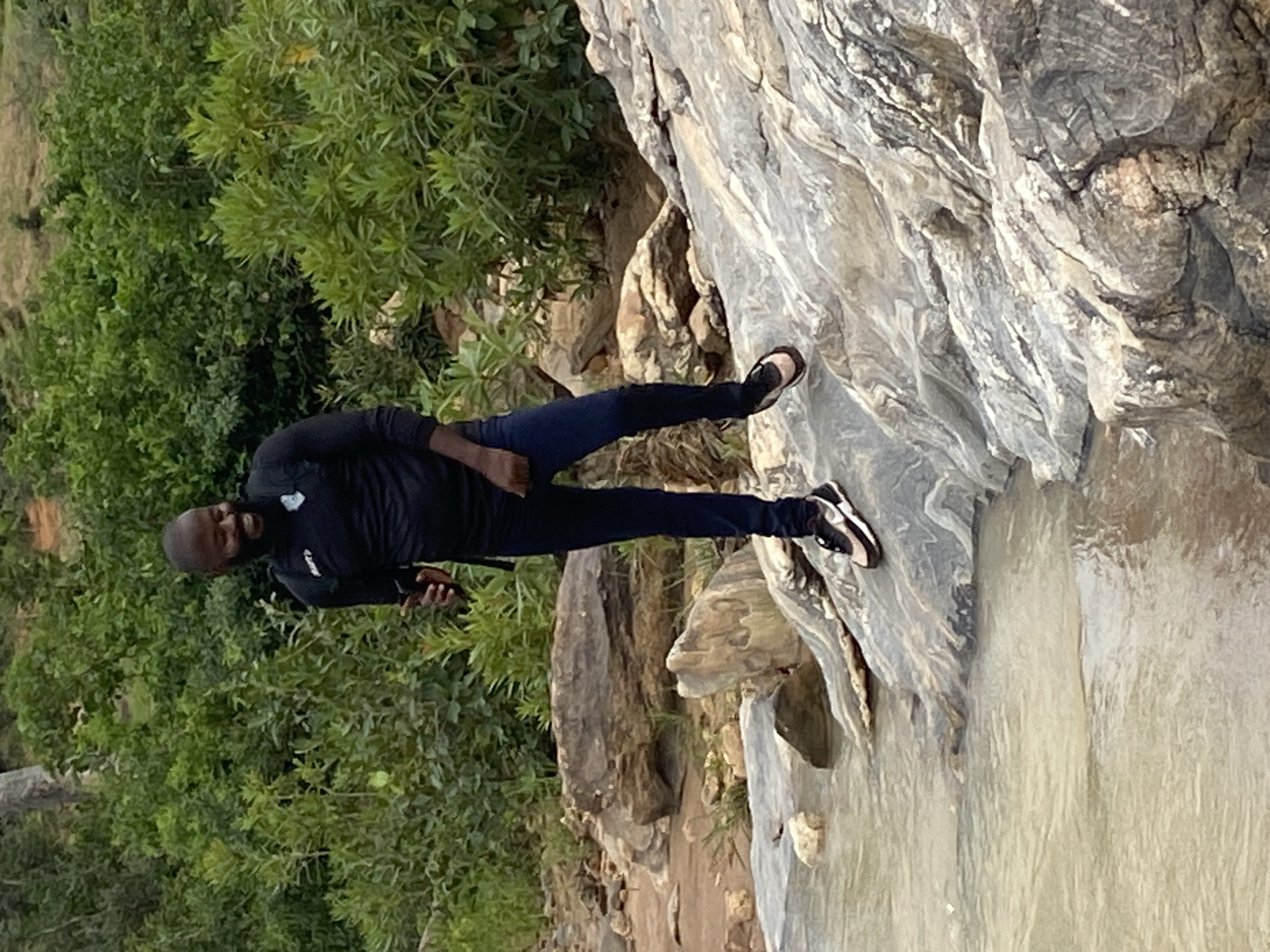 A bald, dark-skinned man wearing a black long-sleeved shirt, dark pants, and black shoes stands on a gray rock formation, facing left. He holds a small black object in his right hand. To his left is green foliage. To his right is a white, flowing stream of water. The location is Ganawuri, Nigeria.