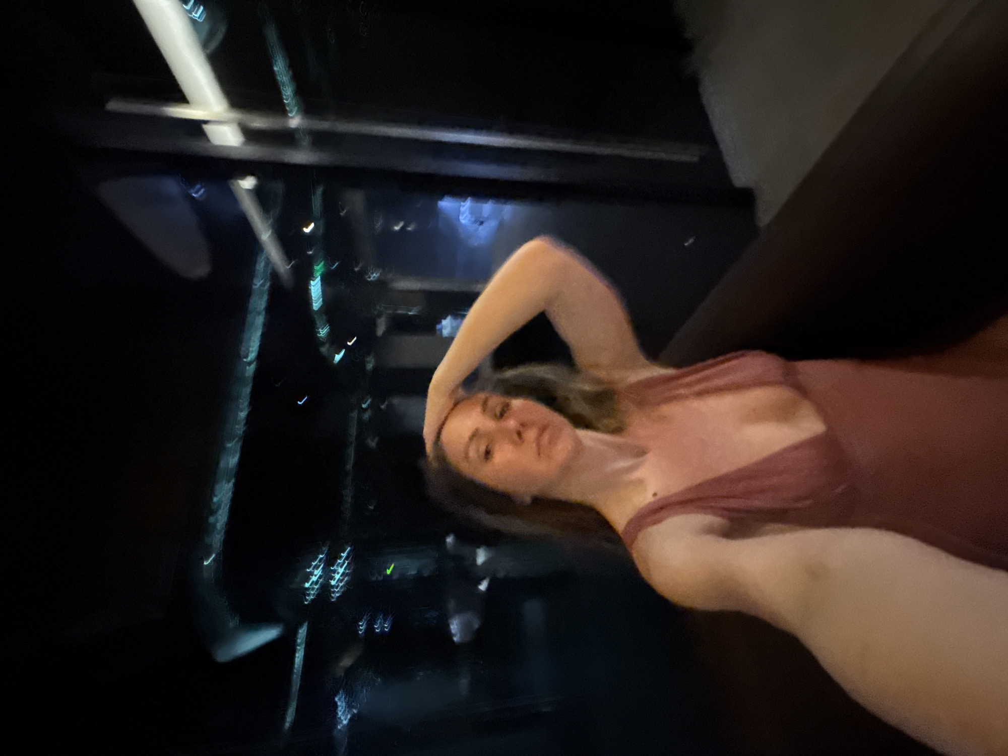 A woman is taking a selfie in a dark environment, likely at night. She is wearing a pink, low-cut dress. Her left arm is raised, with her hand resting on her head. The background is blurry and illuminated by streaks of light, suggesting movement or reflections from city lights or a vehicle. The overall impression is that of a casual, personal moment captured in a dimly lit, possibly urban setting. There is no discernible indication of Sofia, Bulgaria.