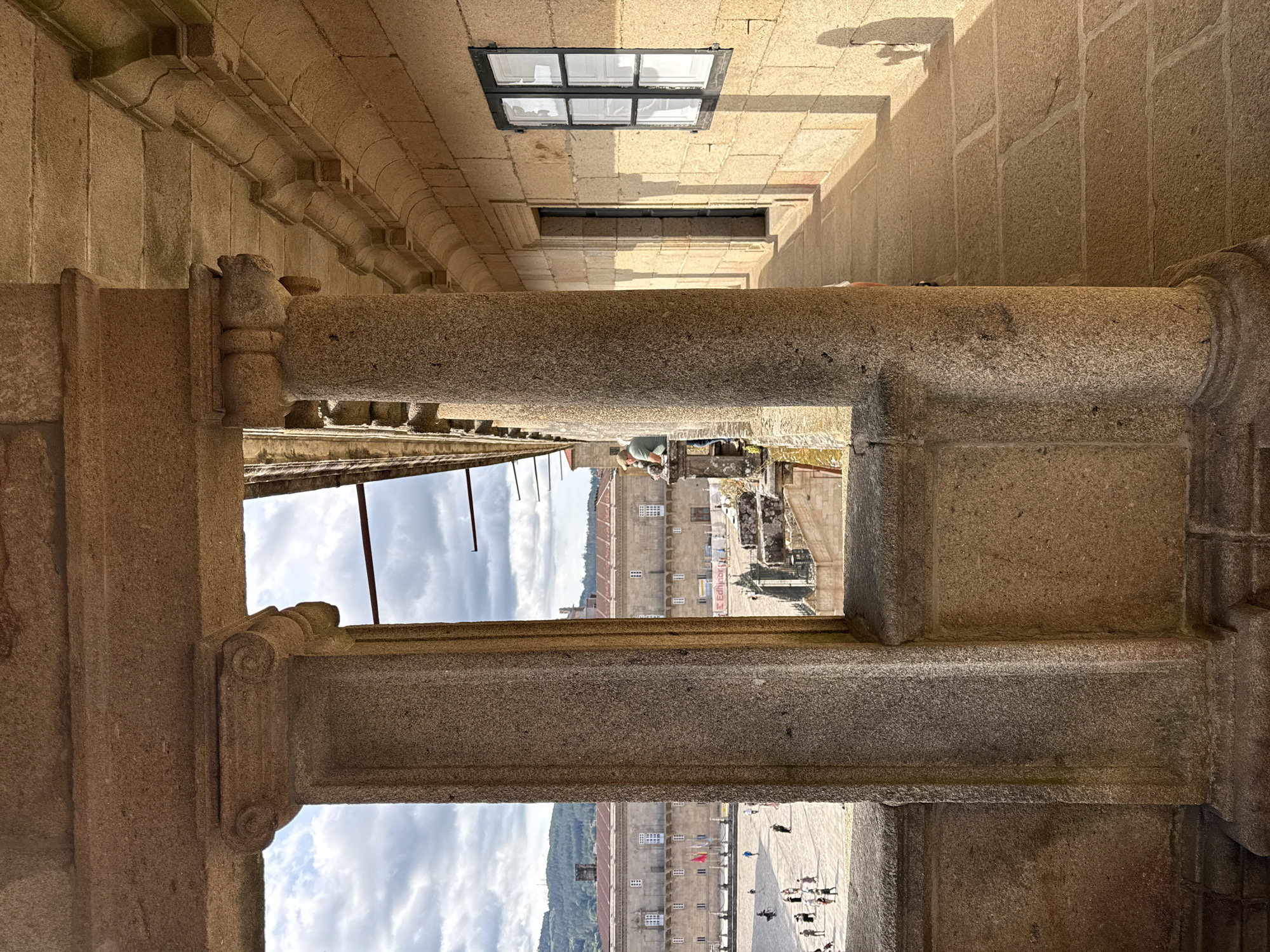 The image depicts a sunlit stone corridor within the Santiago de Compostela Cathedral complex in Santiago de Compostela, Spain. Large, rough-hewn stone pillars frame the view, with the sunlight casting long shadows across the stone-paved walkway. To the left, through an opening in the pillars, a bustling plaza is visible, with numerous pedestrians, flags, and buildings. To the right, the corridor extends into the distance, featuring a stone wall with a small, dark-paned window. A single person, dressed in a green shirt and light-colored pants, leans over a balustrade to the left, looking out at the plaza. The architectural style suggests a historic European religious site.