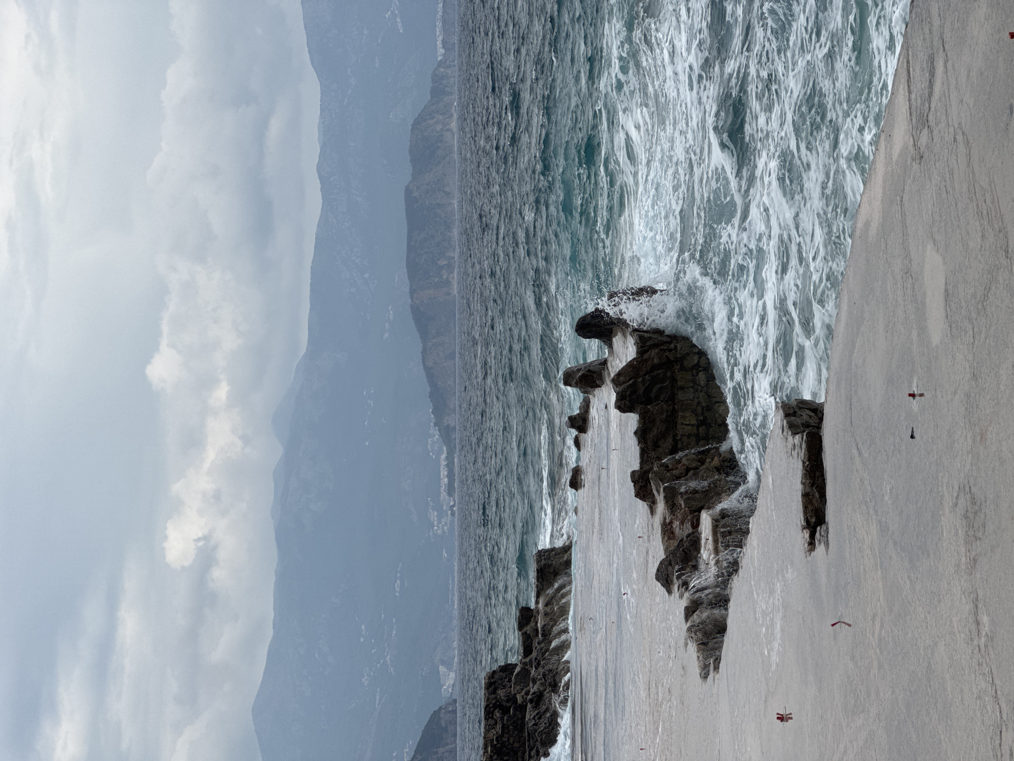 The image captures a dramatic coastal or lakeside scene under an overcast sky, viewed with a horizontal orientation. In the foreground, on the right, a wide, flat, grey surface, possibly a concrete pier or a natural rock platform, extends into the frame. Several small, red, rectangular or L-shaped markers are visible on this surface, scattered intermittently. To the left of this platform, dark, rugged rocks line the water's edge, where powerful waves crash and break, creating white foam and spray.
The central part of the image is dominated by a large body of water, which appears turbulent and choppy with numerous whitecaps, suggesting strong currents or wind. The water exhibits shades of dark blue and grey-green. Across the water, in the background, a series of dark, imposing mountains or steep hills rise, their peaks partially obscured by heavy grey clouds or atmospheric haze, creating a sense of depth and scale.
The sky above is entirely covered with dense, grey and white clouds, indicating an overcast day. The overall impression is one of natural wildness and raw power, with the rough sea and rugged landscape set against a gloomy sky. There are no people visible, and no discernible text appears anywhere in the image. The scene conveys a sense of solitude and the untamed beauty of nature.