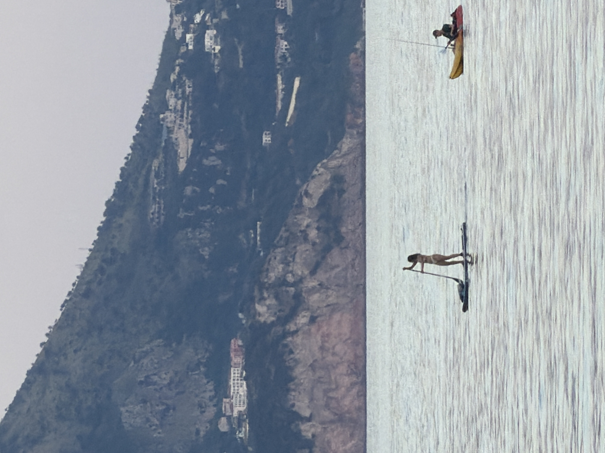This image captures a serene coastal scene in Budva, Montenegro, featuring a steep, verdant mountain rising from the sea under a bright, possibly overcast sky. The mountain is dotted with numerous white and light-colored buildings, indicating settlements or resorts on its slopes.
In the foreground, on the calm, slightly rippled water, two individuals are engaged in recreational activities. On the right, a person is seated in a red and yellow kayak, holding a paddle. To the left, a person is visible performing what appears to be a yoga pose or stretch on a stand-up paddleboard (SUP), with their body facing the left side of the frame and their arms extended forward on the board.
The light suggests it is daytime, possibly morning or late afternoon, with a soft, diffused quality. There is no discernible text visible in the image. The overall impression is one of leisure and natural beauty.