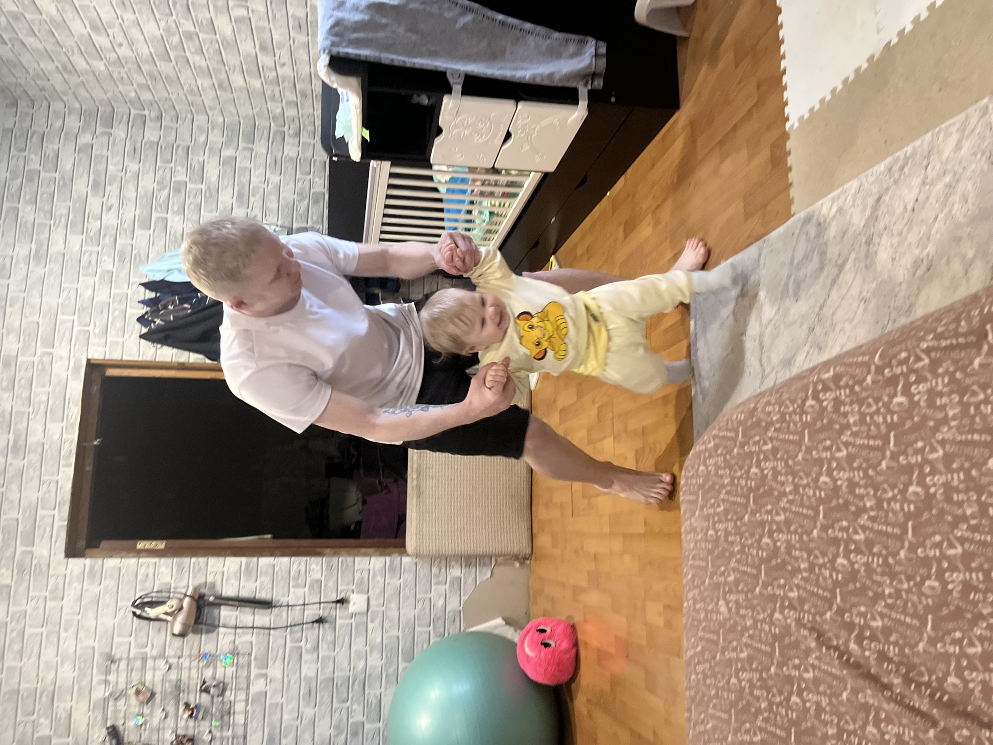 An adult male and a young child are captured indoors, engaged in play. The adult, with light blonde hair, wears a white t-shirt and black shorts, displaying a tattoo on his left forearm. He holds the hands of the child, who is dressed in a yellow long-sleeved top featuring a depiction of Simba from The Lion King, and matching yellow pants. The child appears joyful, with an open-mouthed expression, likely smiling or laughing, as they are held up, seemingly balancing or taking steps.
The scene is a room with light grey faux-brick wallpaper on one wall. The floor is a mix of warm wooden floorboards, a grey patterned rug, and beige and white interlocking foam play mats. In the background, a dark dresser or changing table with white decorative drawer fronts is visible, with a crib just behind it. To the right, part of a bed covered with a brown duvet, patterned with repeating cursive words including "Coffee," is visible. Other items in the room include a large green exercise ball and a bright pink plush toy with a smiley face on the floor. A hairdryer and a small wire grid for displaying pictures are mounted on the wall. A dark, rectangular opening, possibly a window or doorway, is also visible on the left side of the frame. The overall impression is a cozy and active home environment.