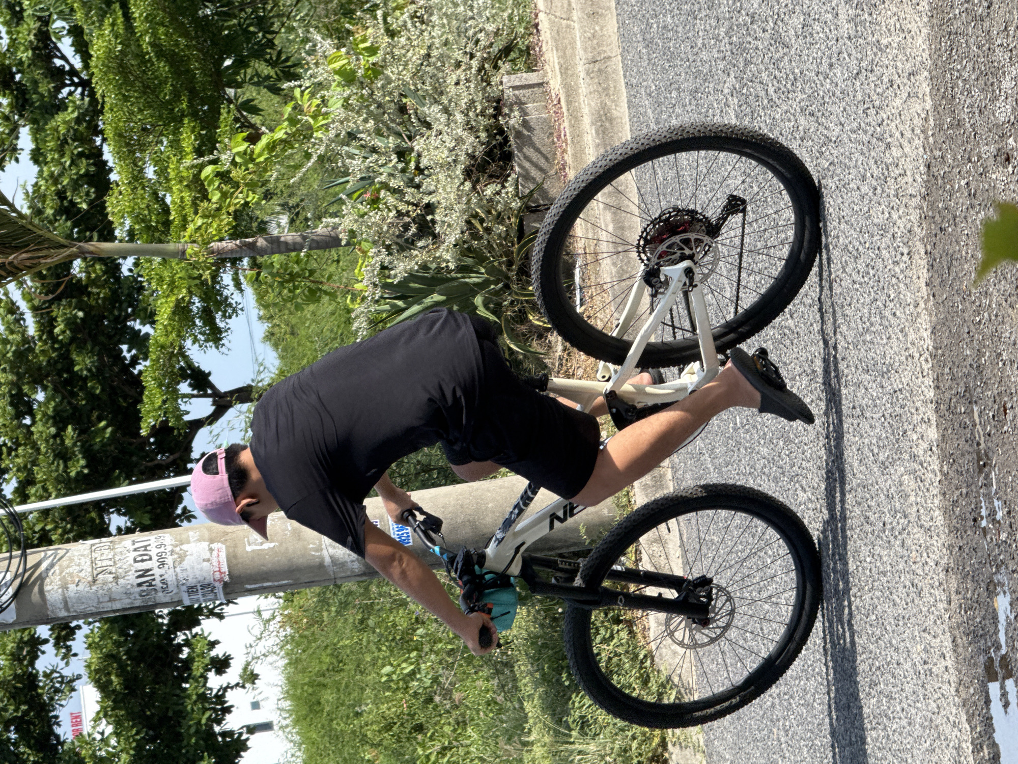 A person wearing a black t-shirt, black shorts, and a pink cap is riding a white bicycle on an asphalt road. The bicycle has a turquoise bag attached to the handlebars. The person is facing away from the camera, and their legs are bent as they pedal. The background consists of a utility pole with several posters attached, lush green trees, and bushes. The image appears to have been taken in a city, possibly Bac Nha Trang Ward, Vietnam, given the presence of Vietnamese text on the posters on the utility pole, including "SAN DAT" and phone numbers. The lighting suggests it is daytime, with shadows cast on the road, indicating sunlight.