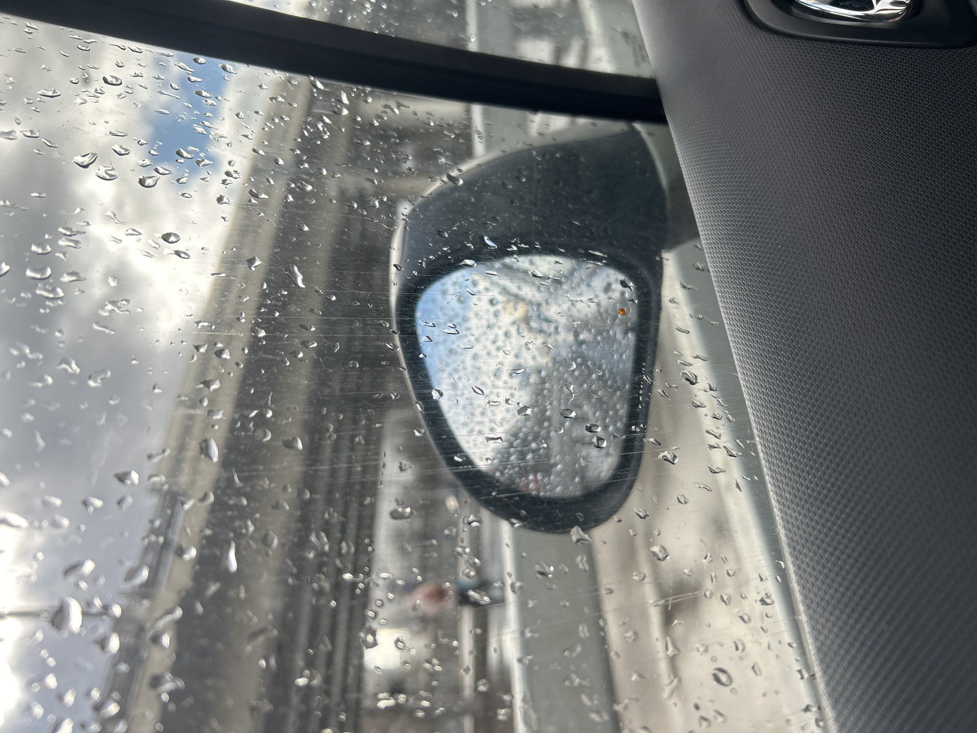 The image shows a view from inside a vehicle, likely a car, through a rain-streaked window. A side mirror is visible, reflecting a blurry scene of a city street. The window is covered in water droplets, obscuring the view. The interior of the car is visible on the right, showing a gray textured surface. The location cannot be determined to be Moscow, Russia with only the given information.