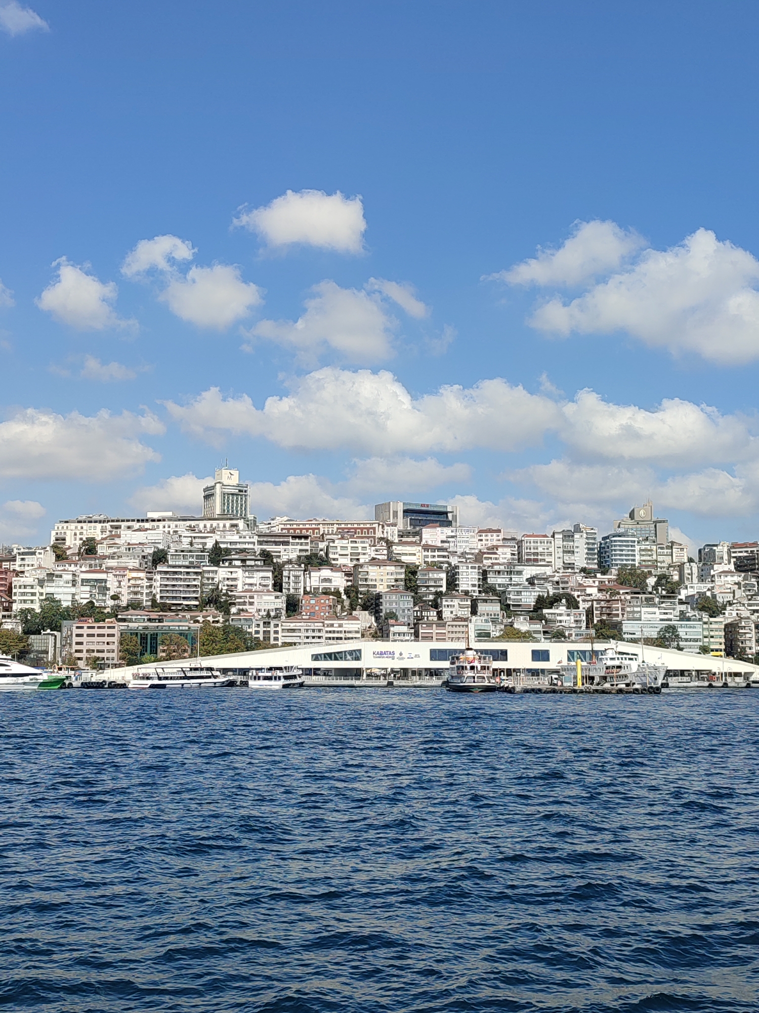 A long shot captures a panoramic view of the Beyoğlu district in Istanbul, Turkey, from across the Bosphorus strait. The image is dominated by a dense cityscape of white buildings with red-tiled roofs, cascading down a hillside. The buildings vary in height and architectural style, with many featuring balconies and large windows.
Prominently visible on the hillside are two distinct modern buildings. One, on the left, is a tall, light-colored building topped with a clock tower. To its right, a slightly shorter, modern building with a curved facade is visible.
Along the waterfront, a long, white modern structure with large windows serves as a ferry terminal. Several boats and ferries are docked at the terminal or in the water in front of it. The largest boat, a white yacht with a green hull, is moored on the far left.
The water in the foreground is a deep blue, with gentle ripples indicating a calm sea. The sky above is a bright blue, dotted with fluffy white clouds. The overall scene depicts a vibrant urban landscape meeting the sea under a clear, sunny sky.