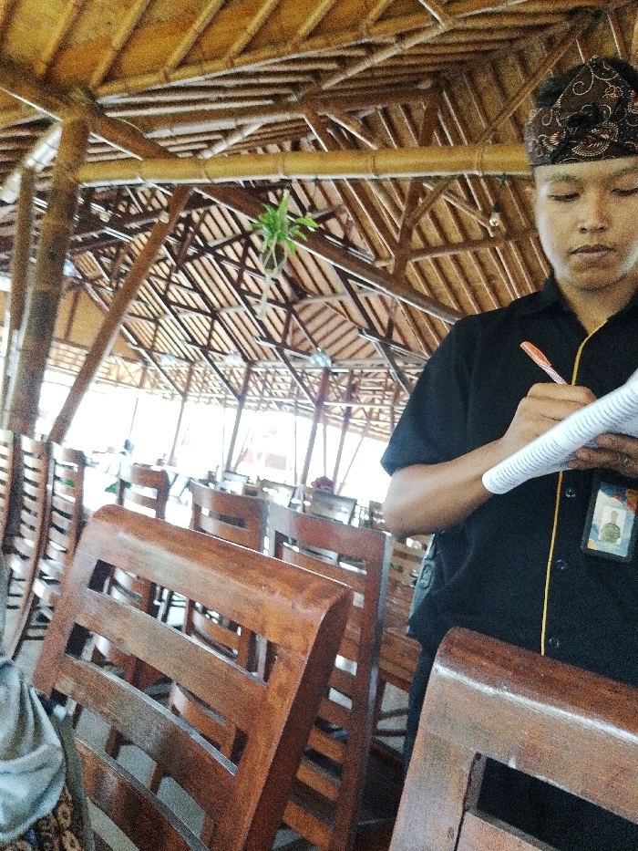 This image captures the interior of a dining establishment in Pabelan, Indonesia. A male individual is visible on the right, from the upper torso, wearing a black short-sleeved shirt with yellow piping, a patterned brown head covering, and an ID badge clipped to his left chest. He holds a white spiral-bound notepad in his left hand and an orange and white pen in his right hand, positioned as if taking notes or an order. His gaze is directed downwards towards the notepad. The background reveals an array of unoccupied dark wooden chairs with slatted backs, arranged in rows. The ceiling and support structure of the building are constructed from numerous interconnected bamboo and wooden beams, forming a visible lattice-work. A small green plant hangs suspended from the overhead structure in the middle-left. The environment is well-lit by natural light, indicating an open-air or semi-open setting.