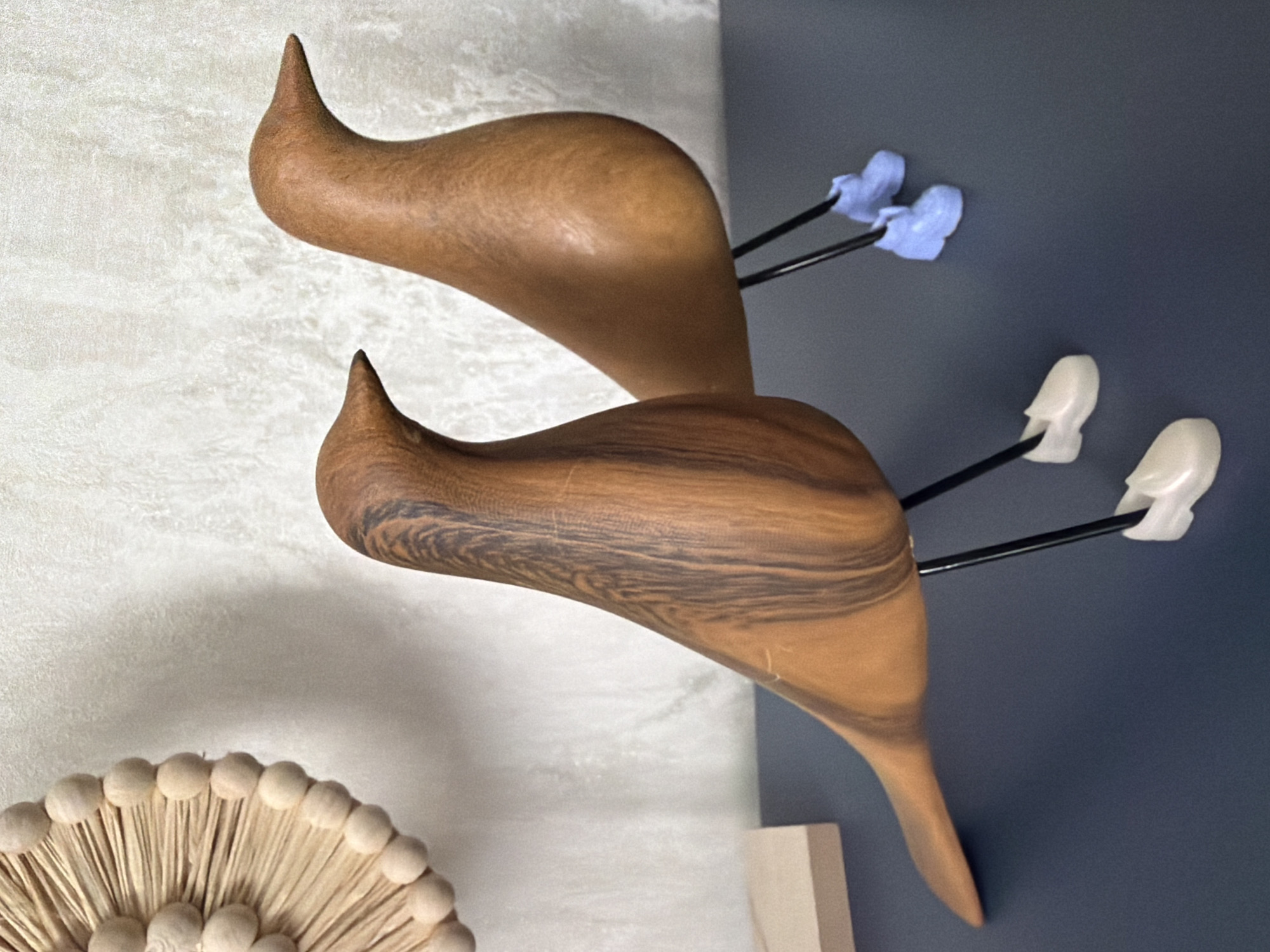 A close-up shot shows two wooden bird figurines with dark vein patterns on a dark blue surface. The figurine in the foreground is on the left, and the second bird is slightly behind and to its right. Both birds have thin black legs. The bird on the left has white shoes, and the bird on the right has light blue shoes. In the upper left corner, a portion of a circular, light-colored wall decoration made of wood beads and straw is visible against a textured white wall. The scene is lit from the front, casting subtle shadows. The image does not provide any information about the city of Novosaratovka, Russia.