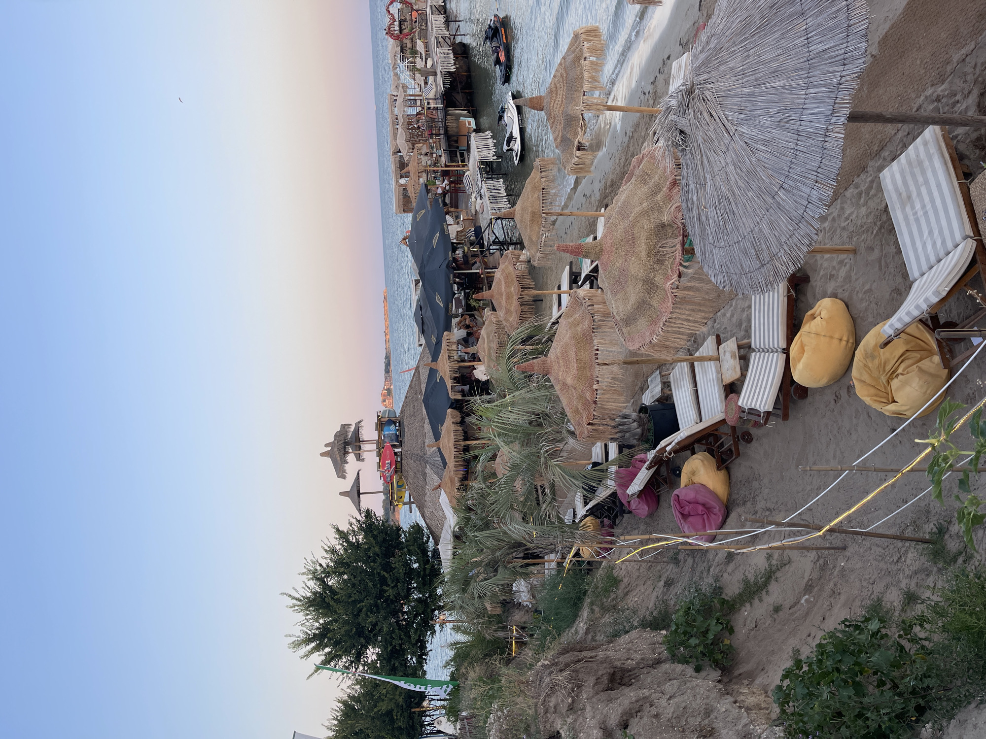 The image depicts a beachside establishment in Ravda, Bulgaria, at dusk. The scene features a sandy beach with numerous sun loungers and straw umbrellas, some with fringed edges. Several beanbag chairs in yellow and pink are scattered amongst the loungers. A pathway made of woven mats leads towards a wooden pier extending into the sea. On the pier, there are more seating areas with umbrellas and people seated at tables. Two jet skis are visible in the water, one black and one white. Palm trees and other greenery are present along the shoreline. The sky transitions from a clear blue at the top to a soft pink and orange hue near the horizon, indicating the setting sun. A single bird is visible in the sky. The overall atmosphere suggests a relaxing beach resort environment.