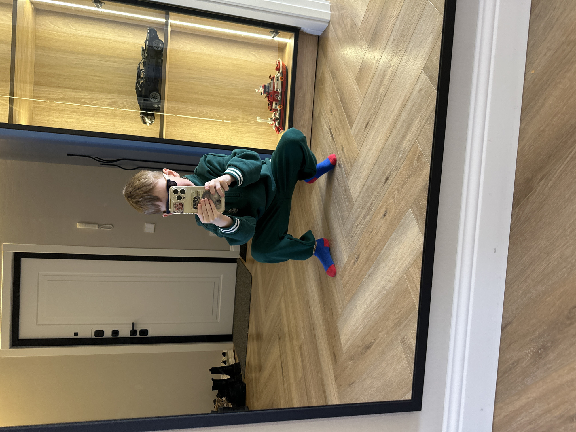 A mirror reflects a boy taking a selfie. The boy is wearing a green tracksuit and mismatched socks (blue with red toes and heels). He's holding a phone with stickers on the case. The reflection shows part of a room with a herringbone wood floor, a display case containing a Lego car and a Lego ship, and a closed white door with black hardware. Beneath the door, several pairs of black shoes are visible. It's impossible to determine the location as Chaikovsky, Russia, without additional information from the image.