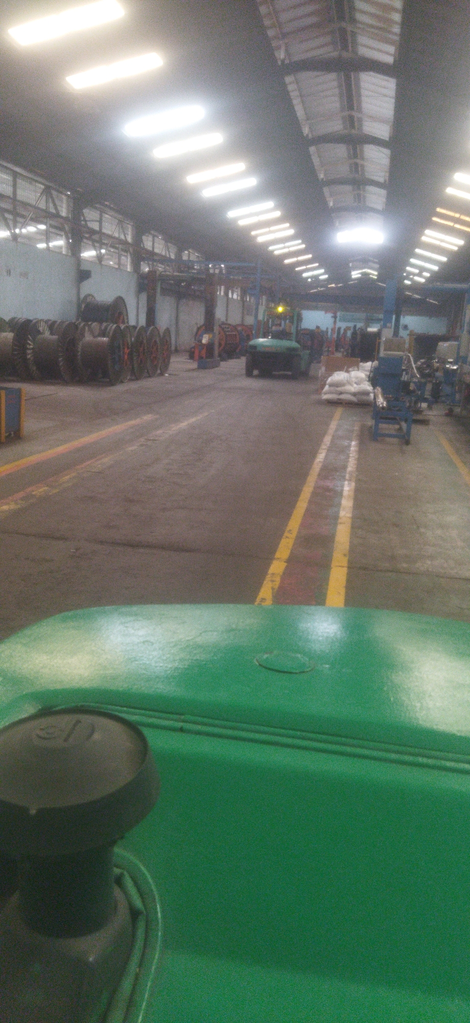 The image depicts the interior of a factory or industrial warehouse, likely located in Cileungsi, Indonesia. The perspective is from the driver's seat of a green vehicle, possibly a tractor or forklift, looking forward. The factory floor is paved and marked with yellow and red lines. To the left, a large collection of spools, likely for wire or cable, are stacked. In the center of the image, a green forklift is visible, moving down the aisle. Beyond the forklift, the factory extends, with more industrial equipment and materials. The ceiling is high, featuring a series of fluorescent lights that illuminate the space. The overall environment appears to be a working industrial facility with signs of ongoing operations.