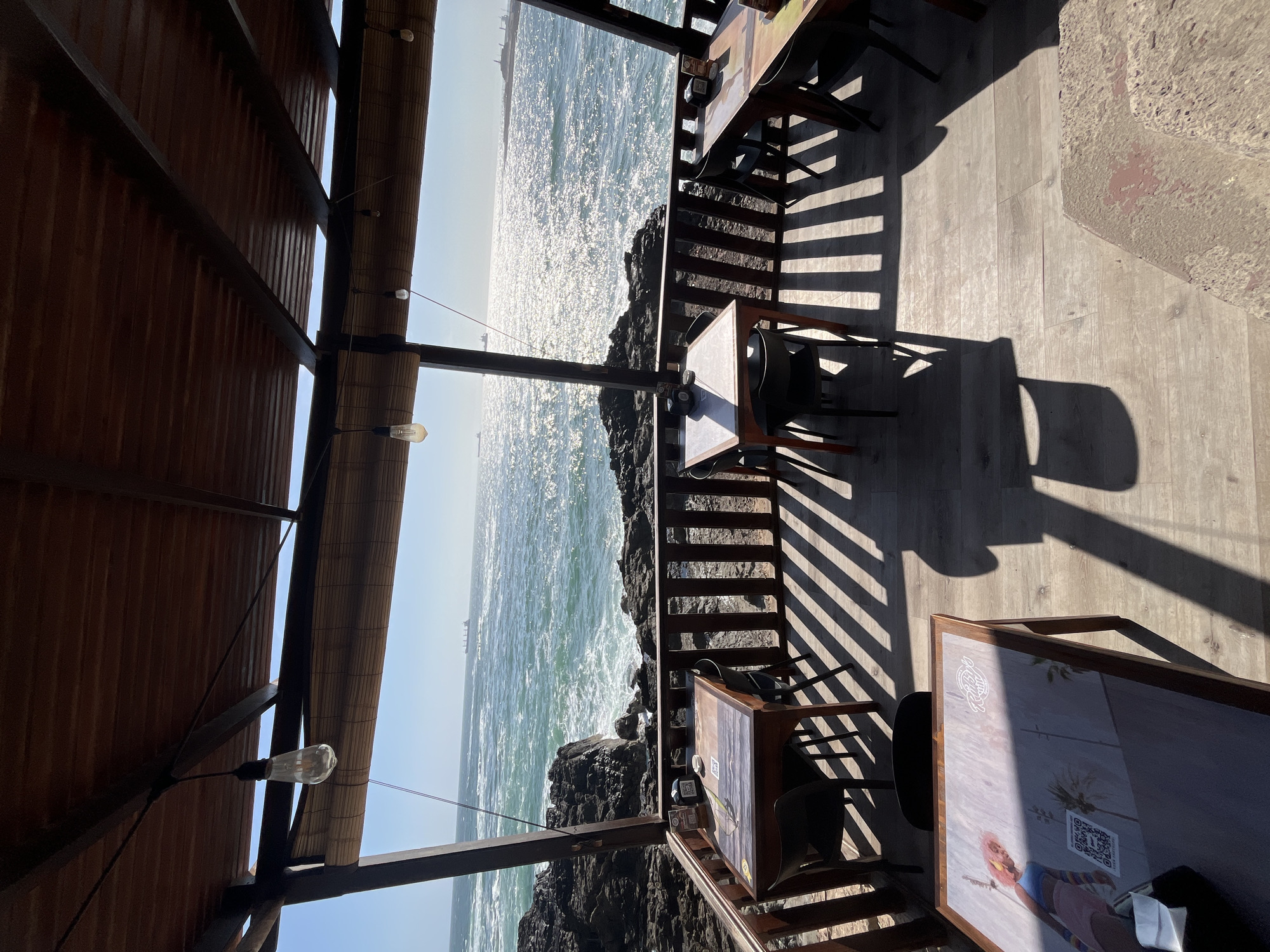 This is an outdoor restaurant scene in Arica, Chile, with a view of the Pacific Ocean. The restaurant has a wooden structure with a slatted roof, and string lights hang from the ceiling. Tables and chairs are set up on a wooden deck. The ocean is visible in the background, with sunlight reflecting off the water. Rocks are visible at the base of the deck, meeting the ocean.