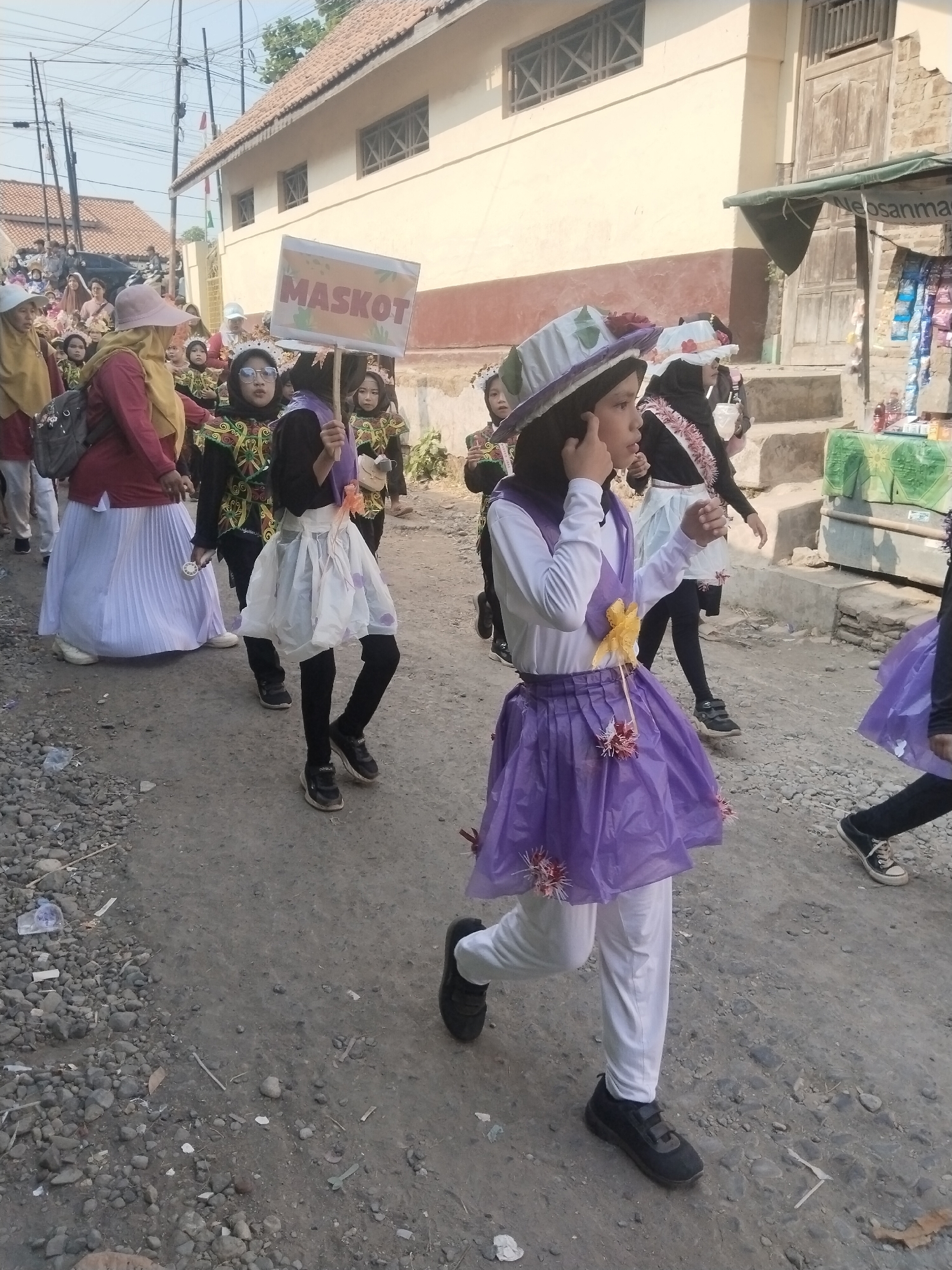 The image depicts a street scene in Jembayat, Indonesia, likely during a parade or procession. A group of young girls dressed in festive costumes are walking down a dirt road. The girl in the foreground, wearing a purple layered skirt and a white top with a yellow embellishment, is looking to the side. She has a dark headscarf and is wearing a hat. Behind her, other girls in similar attire are also participating. One girl holds a sign that reads "MASKOT". The background shows buildings with tiled roofs, a street vendor stall with goods displayed, and utility poles. The road is lined with gravel and some scattered debris. The overall atmosphere suggests a community event.