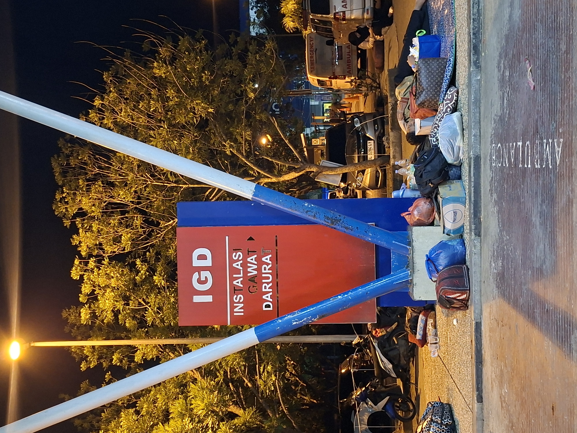 This nighttime image depicts an outdoor scene at the emergency department entrance of a hospital in Banda Aceh, Indonesia. A prominent red and blue sign in the center displays "IGD" in large white letters, followed by "INSTALASI GAWAT DARURAT," indicating "Emergency Department" in Indonesian. This sign is supported by two converging blue and white poles.
On the right side of the frame, an individual is lying prone on a patterned mat with a pillow and blanket, appearing to be asleep, surrounded by various belongings including a dark patterned handbag, a backpack, blue plastic bags, and a cardboard box. Nearby, another individual wearing a dark headscarf and clothing is seated on the ground, looking downwards, positioned next to a white ambulance van.
Multiple vehicles are present. On the right, two white vans are visible, labeled "EMERGENCY" and "AMBULAN" (Ambulance), with a third vehicle partially seen. A silver Honda car with the license plate "BL 1284 LY" is parked behind the sign. Several motorcycles are parked on the left, near the base of the sign.
The area is illuminated by a bright yellow streetlight positioned on the left, casting light on the surrounding trees with dense green and yellow foliage. The ground consists of textured concrete pavement and asphalt. The word "AMBULANCE" is painted in white letters on the asphalt in the foreground. The overall scene suggests a waiting or resting area for individuals outside an emergency medical facility.