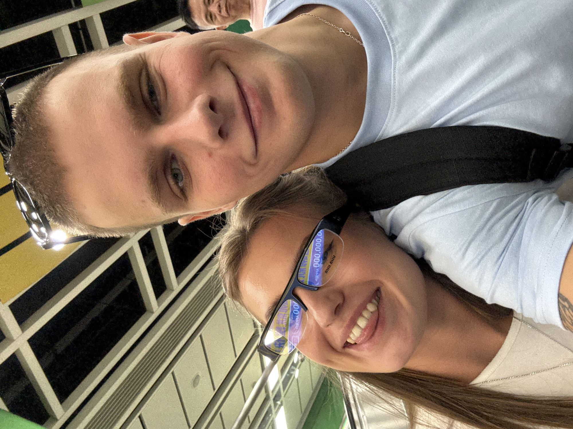 In Haizhu District, China, a young man and woman are taking a selfie indoors. The woman, in the foreground on the left, wears glasses that reflect a digital display with the numbers "000,000.0". She has long brown hair and a white shirt, and is sticking her tongue out playfully. The man, to her right, is wearing a light blue t-shirt and has short brown hair. Behind them, another man with short dark hair is visible in the background. The environment appears to be an indoor public space with a ceiling featuring a pattern of green, yellow, and white panels.