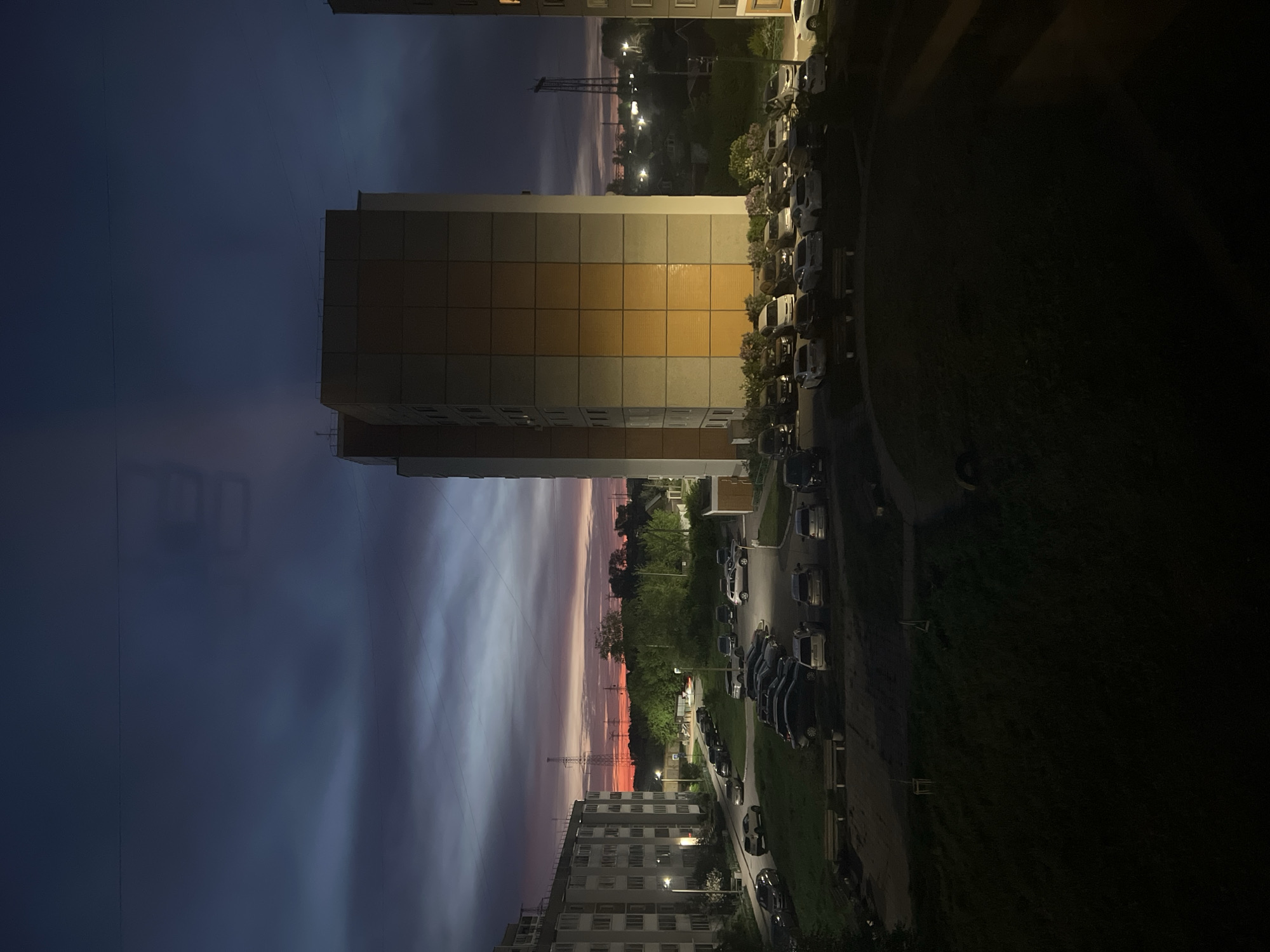 The image depicts an urban scene, likely a residential area in the city of Chaikovsky, Russia, during twilight. A tall, multi-story apartment building dominates the center of the frame, with a distinct color variation on its facade. To the right, a line of parked cars sits alongside a green area. The sky is characterized by a gradient of colors, transitioning from dark blue to shades of pink and orange near the horizon. A transmission tower is visible on the horizon. Another apartment building is on the bottom of the image. The image is oriented vertically.