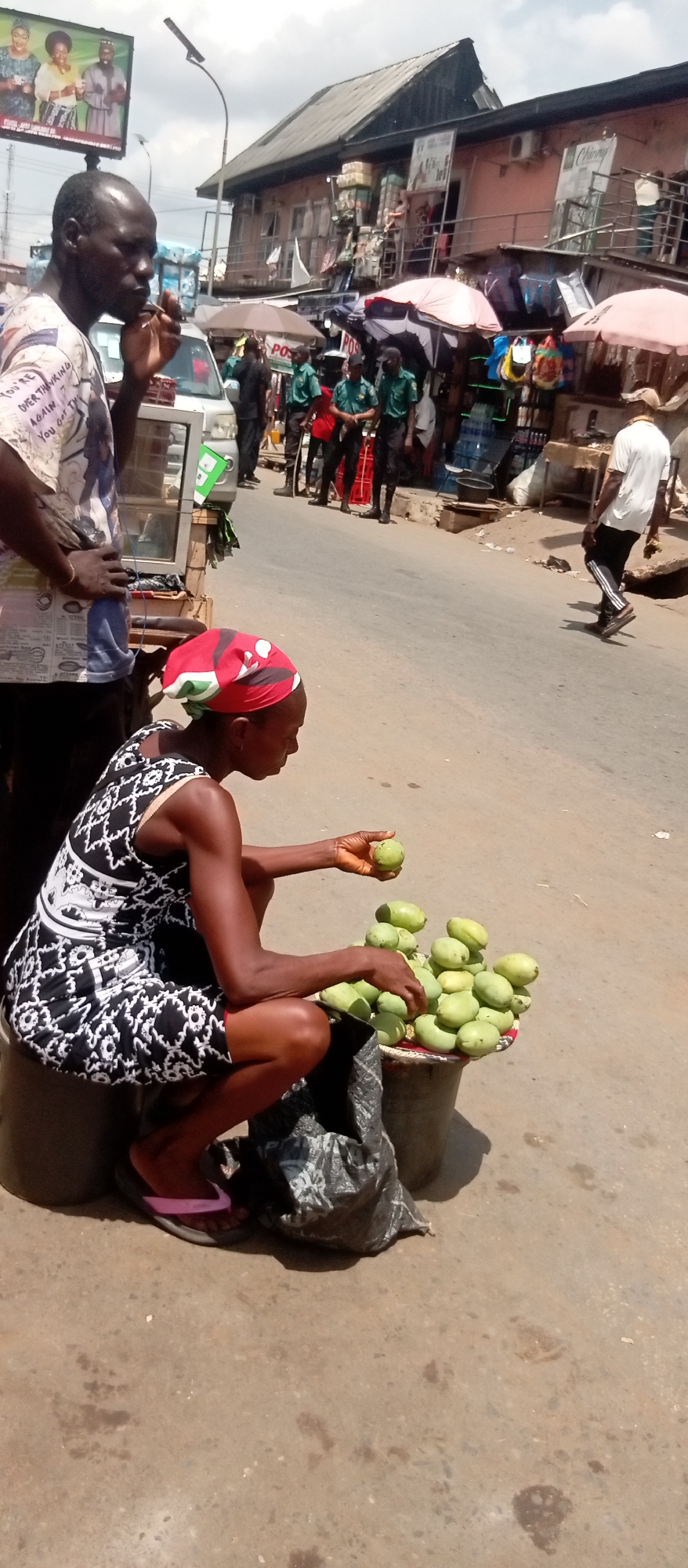 The image captures a vibrant street market scene in what appears to be Onitsha, Nigeria, during daylight hours, likely mid-morning or afternoon, under a partly cloudy sky. The setting is a bustling urban street with buildings lining the roadside, some multi-story, and various stalls and vendors.
In the foreground, a man with dark skin stands on the left, wearing a patterned short-sleeved shirt. Text on his shirt is partially visible, reading "YOU'RE OVER THINKING AGAIN YOU GOT TH...". He holds a small object to his mouth, possibly eating. Next to him, a small glass display case is visible. To the right, a woman with dark skin, wearing a black and white patterned dress and a red headscarf, is crouched low, attending to a display of green fruit, most likely mangoes, arranged on a tray. She holds one fruit in her hand, seemingly sorting or offering them for sale. A black plastic bag rests near the fruit display. She wears pink flip-flops.
The mid-ground features a white vehicle and several individuals. Notably, a group of men in green uniforms and dark pants, possibly security or local officials, stand on the side of the road. Further down the street, a man in a white shirt and dark athletic pants walks away from the viewer.
The background reveals typical market activity with various stalls, large umbrellas providing shade, and merchandise like bottled water. Buildings show signs of commercial use, with signs including "Chianyi" visible on one upper floor. A large billboard featuring people and unreadable text, along with "POST...", is prominent on the left, attached to a street light pole. Another sign with "POST" is visible on a pole in the mid-ground. The street surface is a mix of paved and dirt areas. The overall atmosphere is busy and reflects daily commerce.