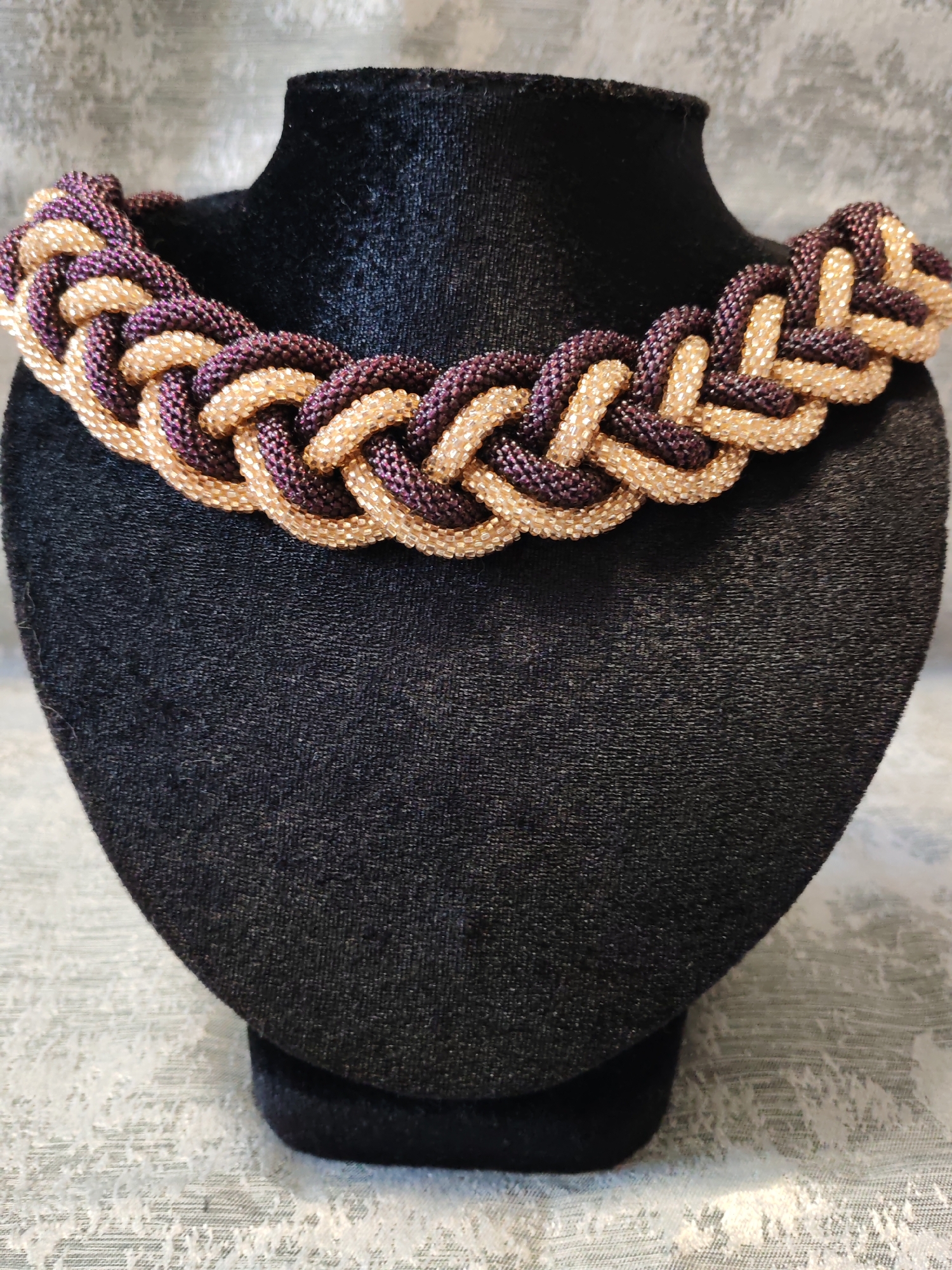 A braided necklace of small gold and purple beads is displayed on a black velvet bust. The background is a light-colored, textured fabric. The image is a close-up shot.