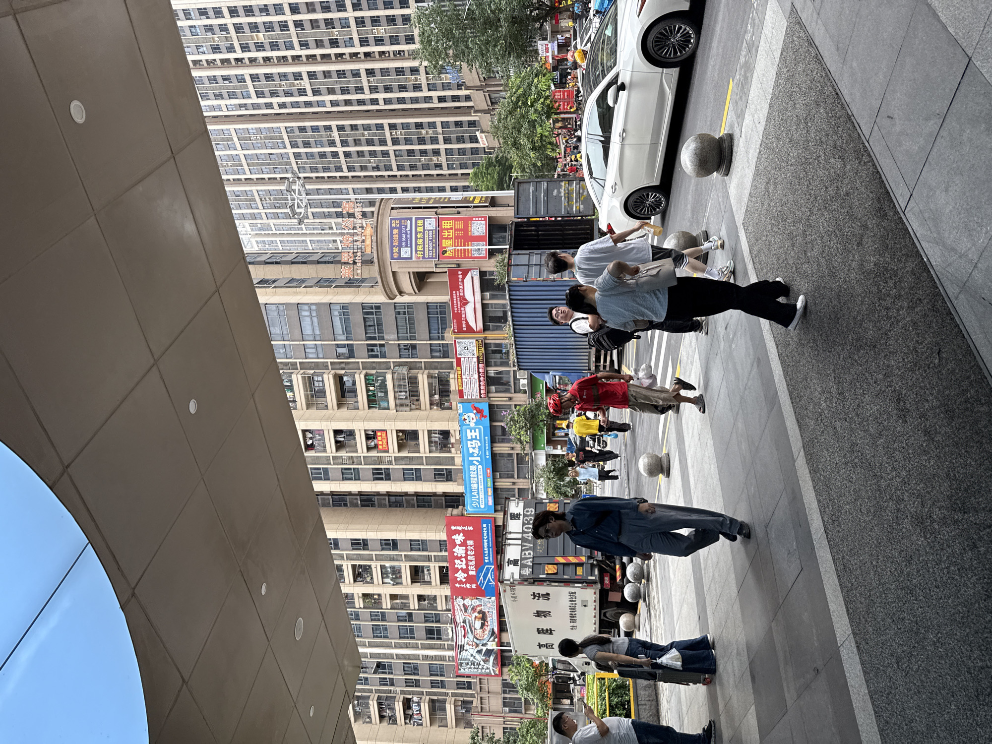 The image depicts a street scene in Haizhu District, China. In the foreground, a gray tiled walkway is occupied by several individuals. A person wearing a dark blue shirt and gray pants walks towards the viewer's left, while another person in a light blue shirt and dark pants walks in the opposite direction. To the right, a white car is parked on the street.
The midground features a large white truck with Chinese characters and the license plate "ABV4039" parked on the street. A blue shipping container is positioned behind the truck, with its doors open. Several pedestrians are walking on the sidewalk.
In the background, tall, multi-story residential buildings dominate the skyline. Billboards with Chinese text are visible on the sides of some buildings. The sky is partly cloudy. The overall impression is one of a bustling urban environment.