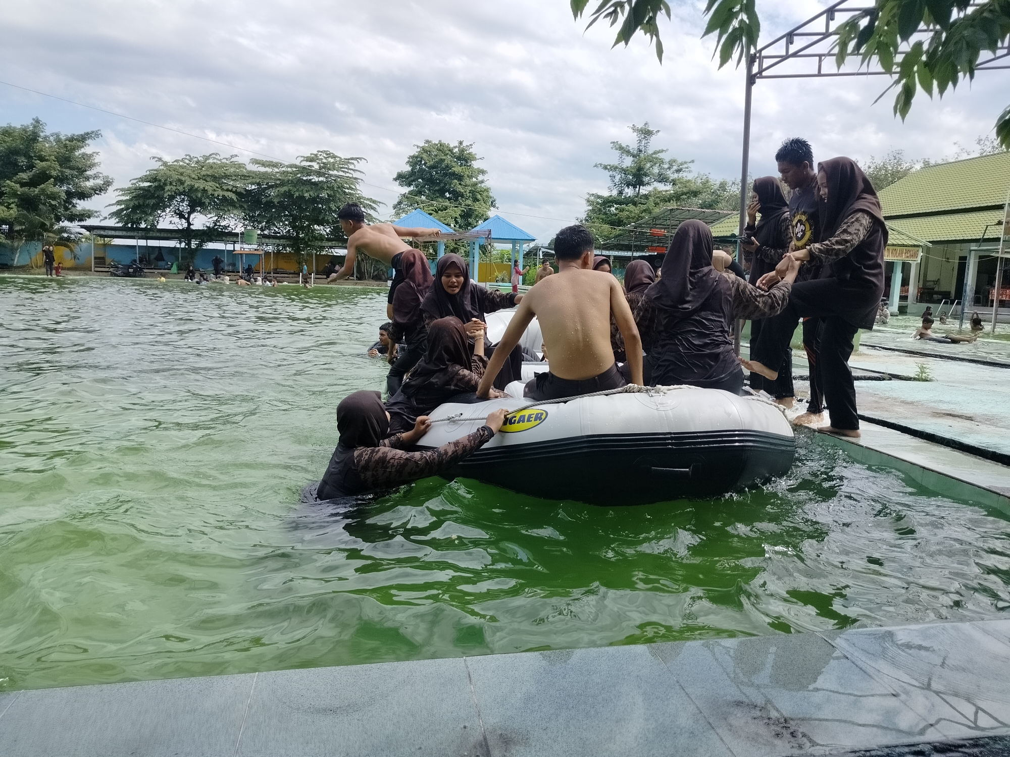 In Tegarmiko, Indonesia, a group of individuals, predominantly young adults, are engaged in water activities at an outdoor swimming pool. The scene captures a dynamic moment where several people are either in the water or interacting with a small inflatable boat. A young man is mid-jump, with his back to the camera, about to enter the water. Others are crowded around the inflatable boat, some inside and others holding onto its sides, appearing to be playing or preparing to move it. The boat itself is white with black trim and has the word "GAER" visible on its side. The water is a greenish hue, and the surrounding environment includes trees, buildings with green roofs, and a cloudy sky. The pool area is bordered by a tiled edge.