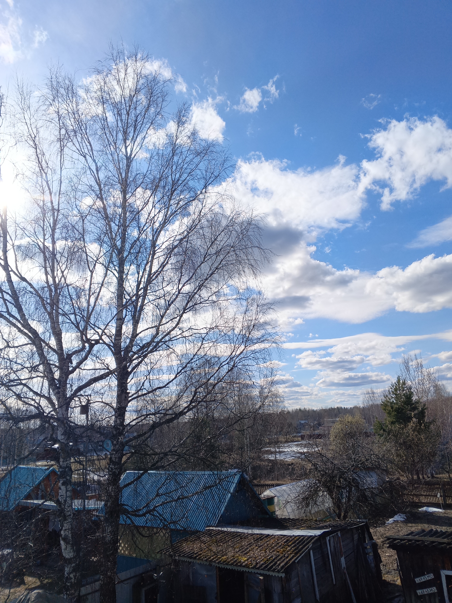 The image depicts a view from an elevated position in Красный Маяк, Russia, showing a landscape with buildings, trees, and a cloudy sky. In the foreground, there are rooftops of various structures, including some with corrugated metal and moss-covered roofs. A large birch tree with bare branches dominates the left side of the frame, partially obscuring the sun. Beyond the buildings, a rural area stretches with a mix of leafless trees, evergreen trees, and patches of snow. The sky is blue with scattered white and gray clouds.
