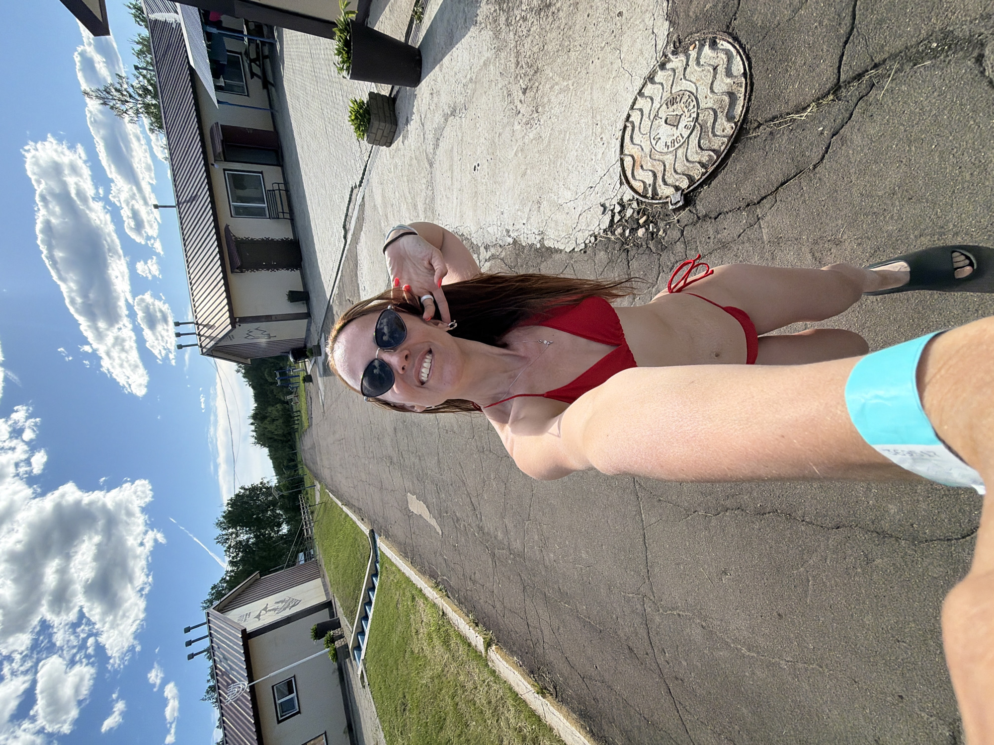 A woman in a red bikini and sunglasses is taking a selfie outdoors. She is standing on a paved road lined with grass and buildings on either side. The buildings appear to be cabins or small houses with brown roofs. The sky is blue with white clouds. There is a manhole cover on the road near the woman. A blue wristband is visible on the arm holding the camera. The overall impression is a resort or vacation setting.
