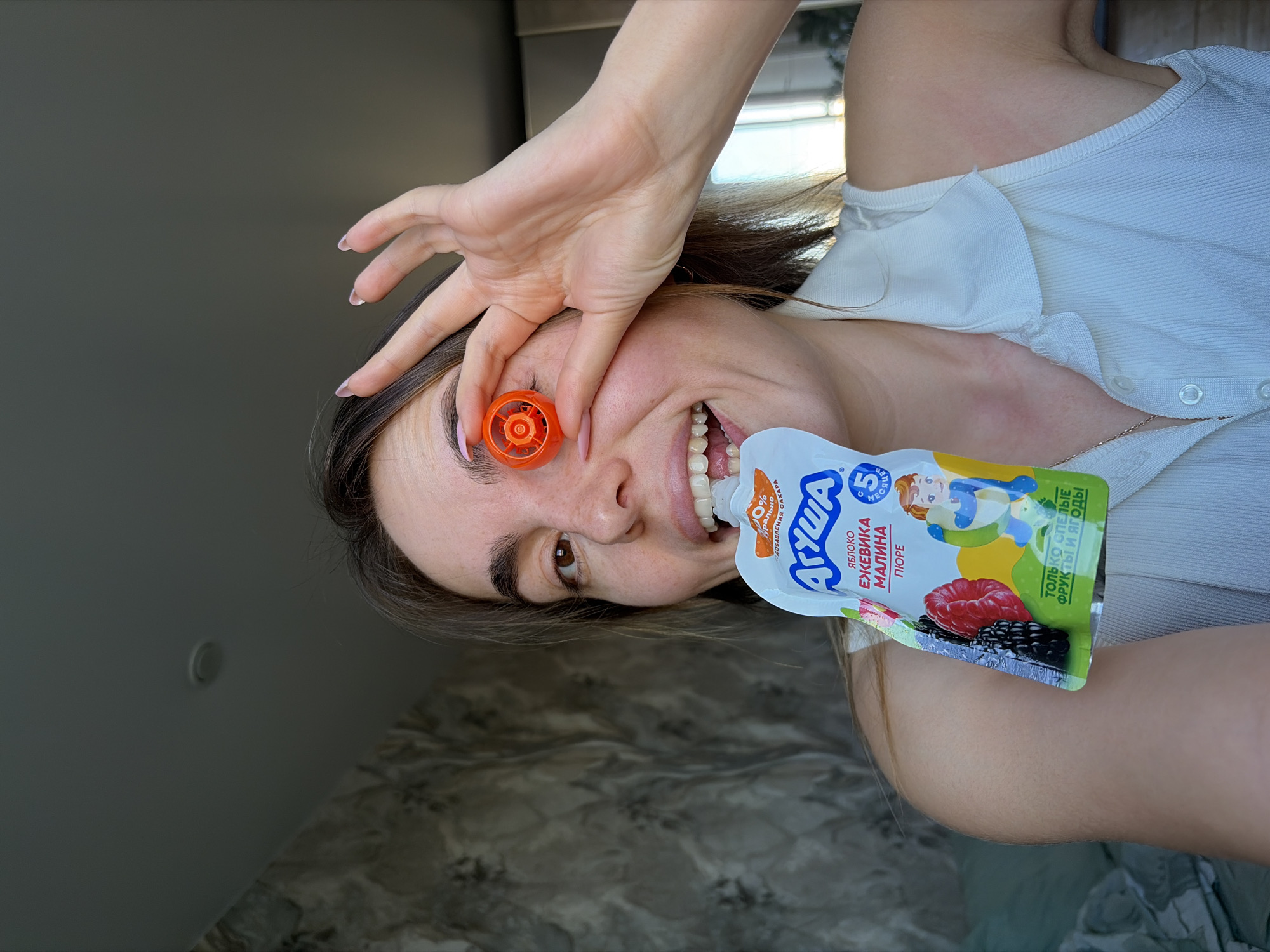 A person is holding a squeezable pouch of baby food in the direction of their mouth with one hand, while the other hand is raised toward their eye with an orange cap obscuring the eye. The pouch is labeled "АГУША" (AGUSHA) and features the text "ЯБЛОКО ЕЖЕВИКА МАЛИНА ПЮРЕ" (APPLE BLACKBERRY RASPBERRY PUREE) indicating the flavor. The packaging also mentions "0% ДОБАВЛЕНИЯ САХАРА" (0% ADDED SUGAR) and "ТОЛЬКО СПЕЛЫЕ ФРУКТЫ И ЯГОДЫ" (ONLY RIPE FRUITS AND BERRIES). The text "5 МЕСЯЦЕ" can also be seen, which translates to "5 Months" - indicating the baby food is suitable for babies 5 months and older.
There's no indication of the location being Izhevsk, Russia.