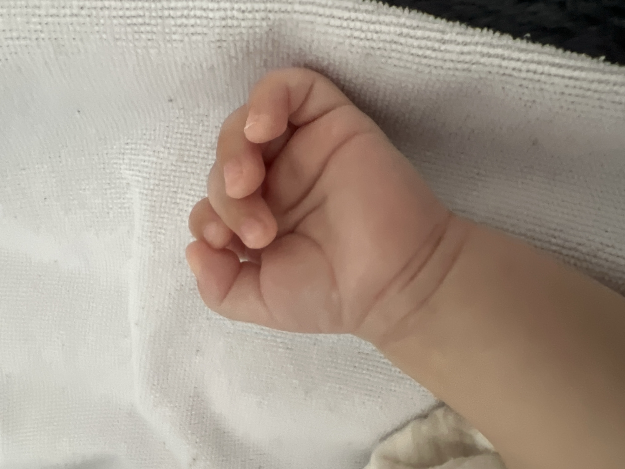 A close-up, overhead shot shows a small, fair-skinned baby's hand with fingers curled into a fist. The hand rests on a textured, light-colored surface, possibly a towel or blanket. The lighting is soft, casting faint shadows that highlight the creases in the baby's skin. No specific location in Gdańsk, Poland, can be determined from the image.
