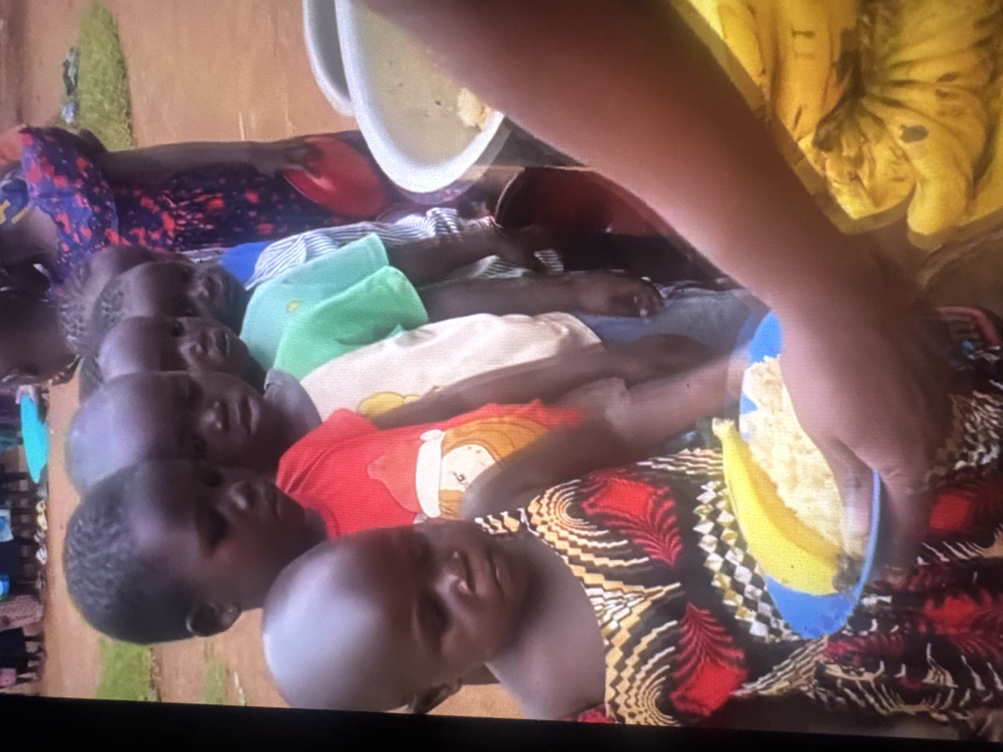 In an outdoor setting, possibly in Abuja, Nigeria, a group of children are lined up, appearing to receive food. In the foreground, a child in a colorful dress is holding a blue plate with a banana and what appears to be rice or porridge. To the right, a person's arm is visible, holding a large bunch of bananas and a white bowl containing food. The children are dark-skinned with short hair, and some are wearing t-shirts with printed designs. The ground appears to be dirt. The overall impression is a scene of food distribution.