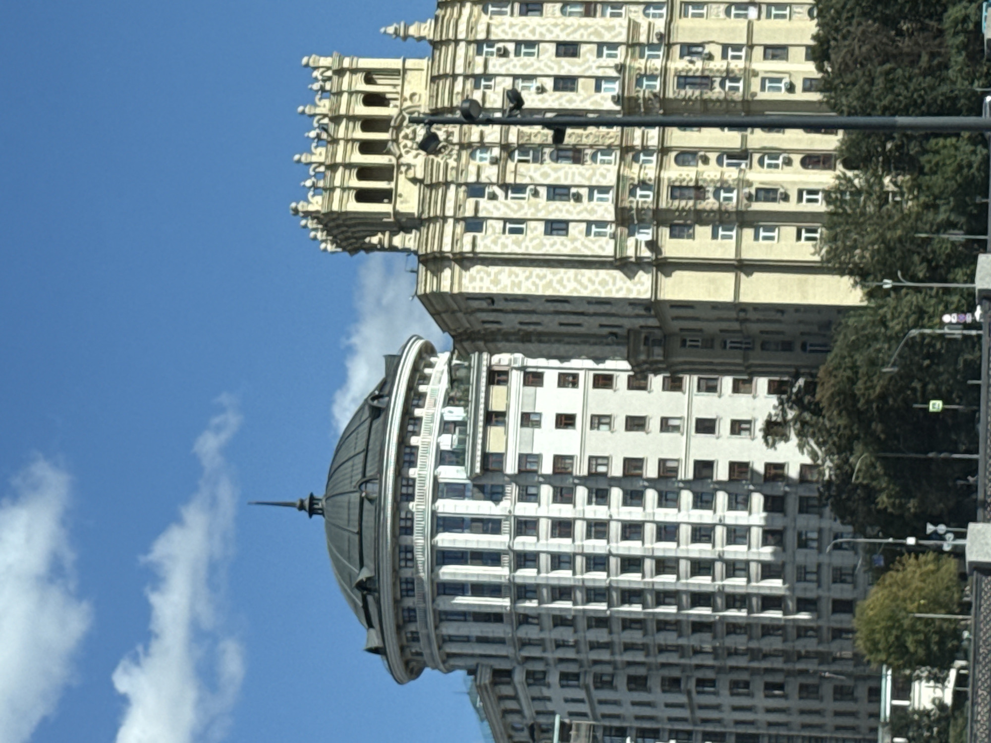 Two tall buildings stand side-by-side under a bright blue sky with scattered white clouds. The building on the left is a cream-colored, rounded structure topped with a dark, domed roof and a spire. It features numerous windows arranged in vertical rows and decorative columns. The building on the right is a taller, yellow, rectangular structure with an ornate, crenellated crown. It also has many windows, some with arched tops, and decorative architectural details. A dark lamppost with multiple lights is positioned in front of the right building. Lush green trees are visible at the base of both buildings, and a body of water or riverfront is suggested by structures at the very bottom. This scene depicts the Stalinist skyscrapers, specifically the Kotelnicheskaya Embankment Building and the one next to it, located in Moscow, Russia, along the Moskva River.