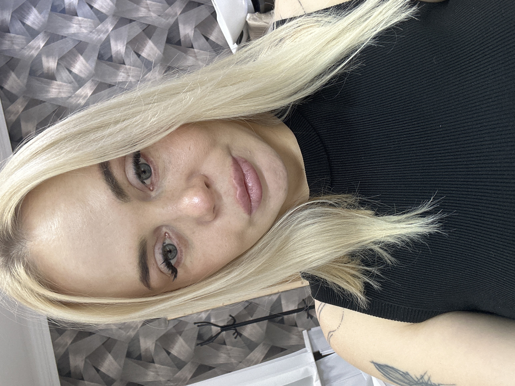 A blonde female is depicted in a close-up, head-and-shoulders self-portrait, looking directly forward. She has long, straight blonde hair with a visible center part, light-colored eyes with apparent lash extensions on the viewer's left eye, and defined eyebrows. Her lips are slightly parted, and her skin shows visible texture, including fine lines on the forehead. She is wearing a black, ribbed, high-neck sleeveless top. Tattoos are visible on her left shoulder and upper arm, consisting of a thin curved line and a stylized feather design. The indoor background features a wall adorned with a three-dimensional geometric pattern composed of interwoven grey and black rectangular or trapezoidal segments. A dark, multi-branched object, possibly a coat stand, is partially visible on the left, alongside a vertical white panel. A white fixture is partially visible on the right. The scene is illuminated by uniform indoor lighting. This occurs in the city of Unknown, Unknown.