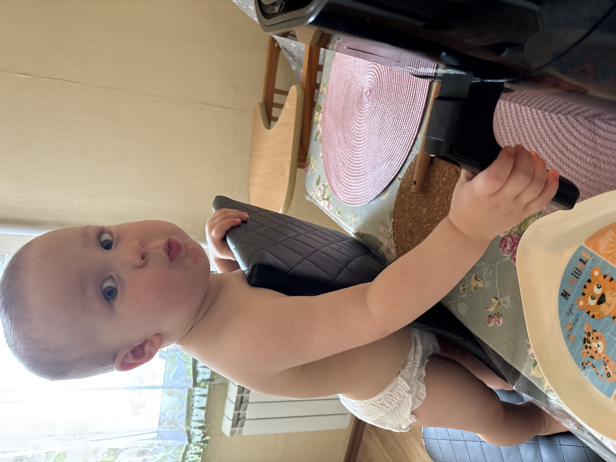 An infant with a diaper is standing and holding onto a black chair. The infant is looking at the camera with a surprised expression. The infant is in a room with a window, a radiator, and a table. There is also an air fryer on the table. The lighting suggests the photo was taken indoors during the day, possibly in Naberezhnye Chelny, Russia, based on the information provided.