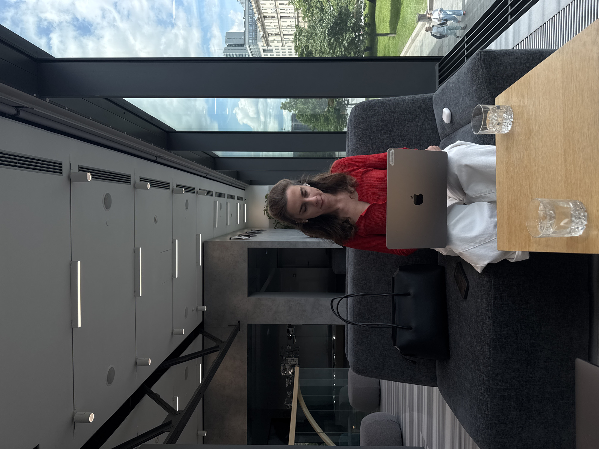 A woman is sitting on a gray sofa, working on a laptop next to a table with two drinking glasses. She is wearing a red sweater and white pants. A black purse is near her on the sofa. The background features a window with a view of buildings and trees, possibly in an urban setting. The building's interior design has white walls with light fixtures. The location is likely a public space such as a lounge or office, in Moscow, Russia.