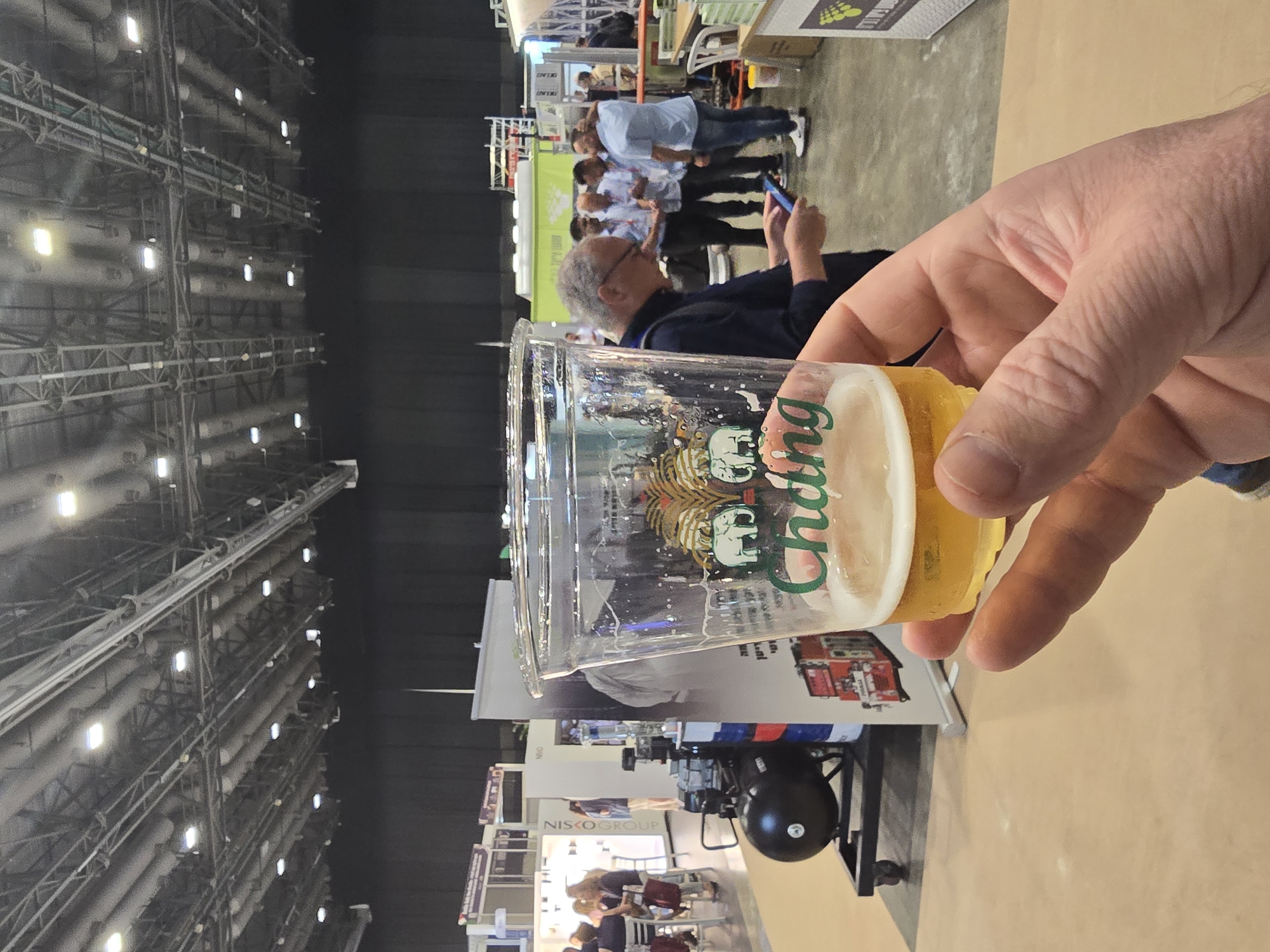 A hand holds a cup of Chang beer in what appears to be a large convention center or trade show in Tel Aviv, Israel. The cup partially obscures the view, which includes trade show booths and people in the background. The convention center has a high ceiling with exposed structures.