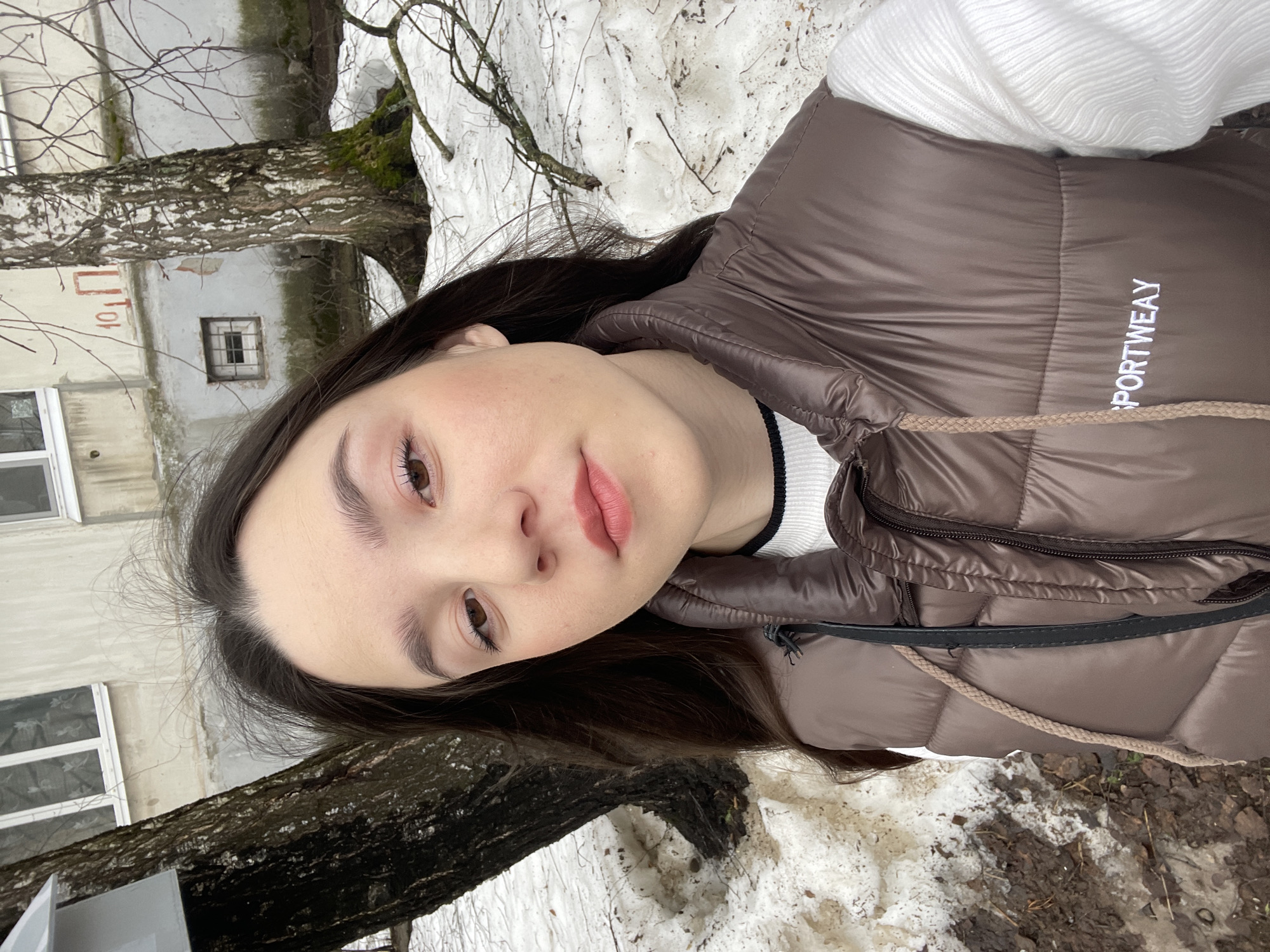 A young woman poses for a selfie in Chaikovsky, Russia. Patches of snow remain on the ground, and a birch tree stands behind her to the left. A building with white-framed windows is visible in the background. The woman is wearing a brown puffer vest over a white sweater with a black neckline. The word "SPORTWEAY" is printed in white on the vest.