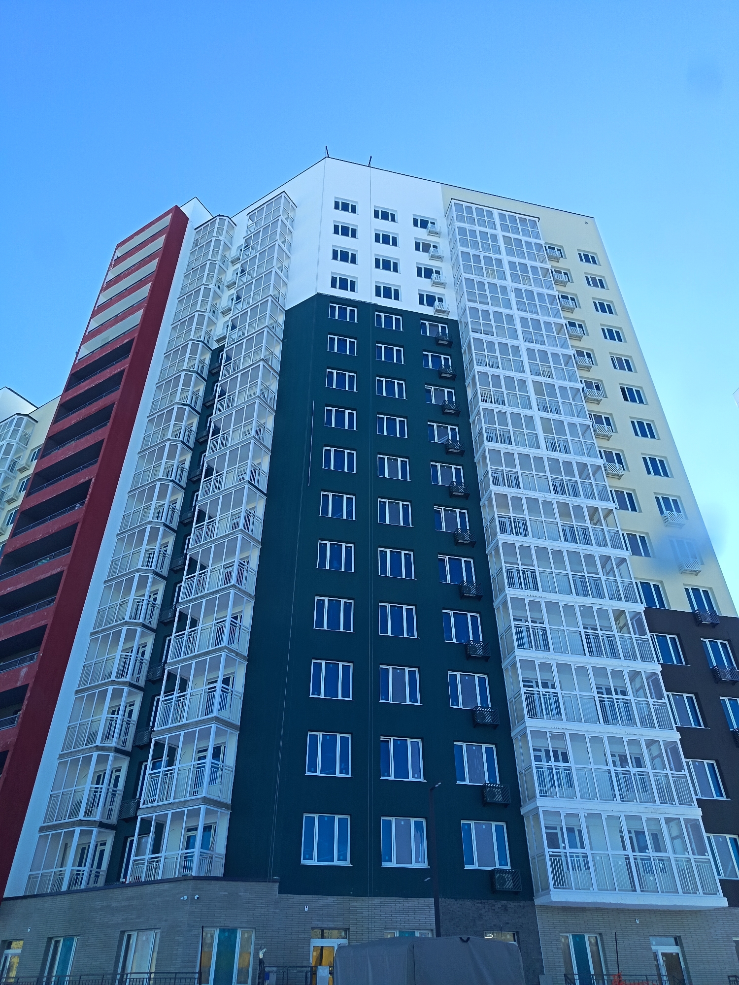 A low-angle, eye-level shot captures a modern, multi-story residential building stretching upwards into a clear, bright blue sky. The building exhibits a contemporary architectural style with a distinctive, colorful facade segmented vertically into several panels.
The leftmost section of the building is a deep red or maroon, featuring horizontal light grey bands that suggest open balconies or fire escapes. Adjacent to this, a broad white section is adorned with numerous glass-enclosed balconies (loggias), extending outwards and creating a textured surface. The central portion of the building showcases a dark teal or blue facade with evenly spaced rectangular windows, primarily with white frames. Moving further right, another white section with glass-enclosed balconies transitions into a light yellow or beige section, also with regular windows. The rightmost visible part of the building features a dark brown or black facade, again with white-framed windows.
Several external air conditioning units are mounted below windows on the darker sections of the facade. The base of the building, partially visible at the bottom of the frame, appears to be clad in a greyish, possibly brick-like material, with larger ground-floor windows or entryways.
The scene depicts the exterior of a newly constructed or recently renovated residential complex, indicative of modern urban development. The time of day is daytime, under clear weather conditions, given the crisp light and shadow definition and the unblemished blue sky. There are no people visible, nor is there any discernible text on the building or in the surrounding environment. The overall impression is one of a functional, geometrically striking structure within an urban setting in Nizhnevartovsk, Russia.