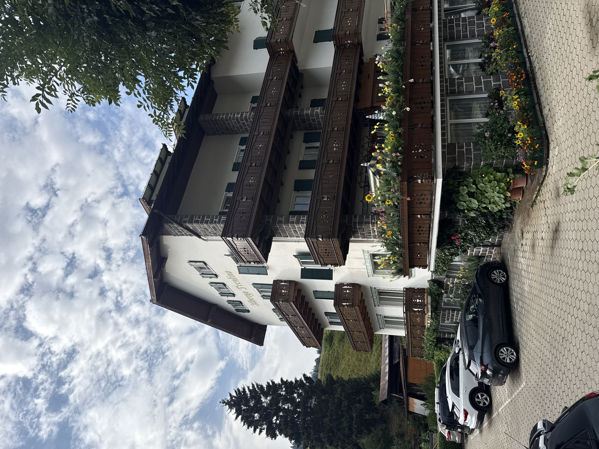 The image depicts the "Albergo Trentino" hotel in Moena, Italy, situated in a mountainous region, likely the Dolomites. The hotel is a multi-story building with a white facade, dark brown trim, and numerous balconies with intricate wooden railings, many of which are adorned with colorful flowers. The building has several windows with green shutters. In front of the hotel, a parking lot is visible, paved with hexagonal bricks. Several cars are parked, including a dark gray hatchback, a white SUV, and a black SUV. The environment surrounding the hotel includes a steep, green hillside, a tall evergreen tree, and some smaller buildings in the background. The sky is partly cloudy with a mix of blue sky and white clouds. The scene appears to be a daytime outdoor shot, possibly during the summer or early autumn due to the blooming flowers. There are no people visible in the image.
