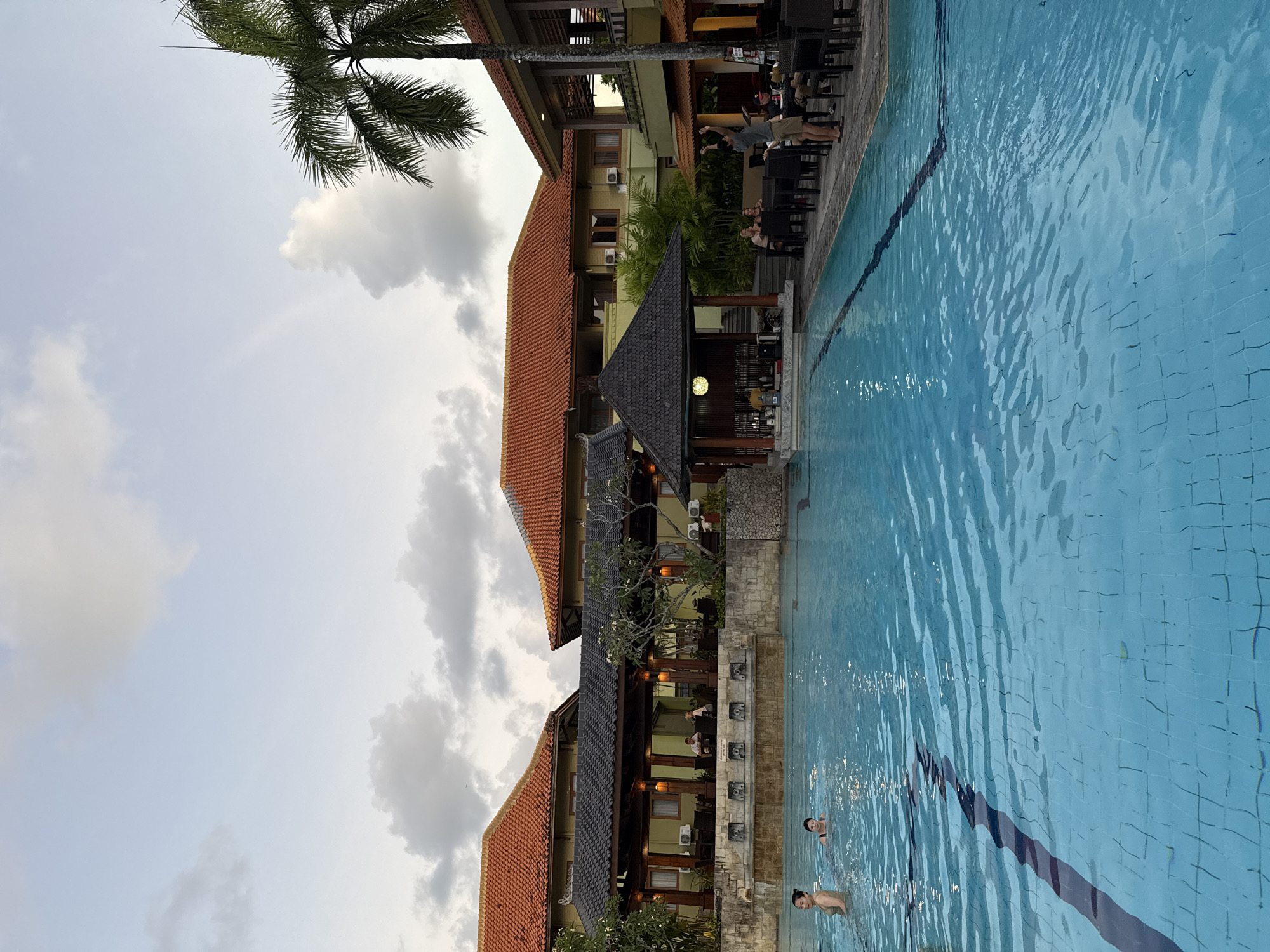 The image captures a vibrant outdoor scene at a resort or hotel in Kuta Selatan, Indonesia, featuring a swimming pool and multi-story buildings under an overcast sky. The bright blue swimming pool dominates the right side of the frame, its tiled bottom clearly visible through the rippling water. At least two individuals are seen swimming or wading in the pool, with one person's head and shoulders visible in the foreground and another person further back.
On the left side of the pool, beyond a low stone wall, stand several interconnected hotel buildings with distinct red and dark-tiled roofs, characteristic of traditional Indonesian architecture. The buildings feature wooden elements, balconies, and air conditioning units mounted on their facades. An outdoor dining or lounge area is visible at the ground level of one building, with numerous tables and chairs. Several people are seated at these tables, appearing to be dining or socializing, with one person clearly standing and stretching.
Lush green foliage, including a prominent palm tree in the upper left corner and other plants surrounding the buildings, adds to the tropical ambiance. The sky above is cloudy and light grey, suggesting either an overcast day or late afternoon. The overall impression is one of relaxation and leisure at a tropical destination. No discernable text is visible in the image.