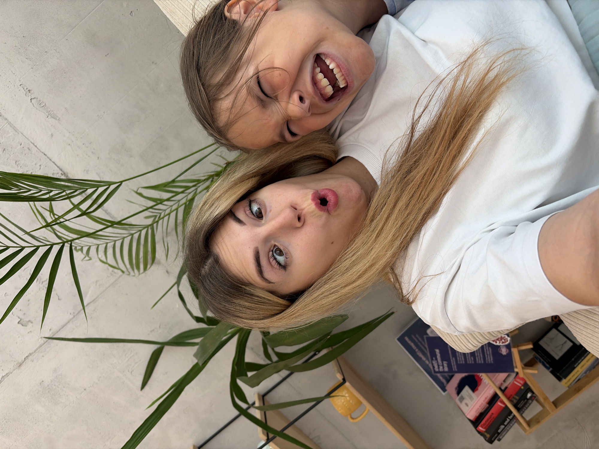 A woman and a child are taking a selfie in an indoor environment. The woman, with long blonde hair, is looking at the camera with wide eyes and her mouth open in a surprised expression. The child, with brown hair, is squishing their face, eyes closed, and mouth open in a wide smile, showing their teeth. Both are wearing white t-shirts. Behind them, a large green plant with fronds is visible against a textured wall. To the left, a wooden shelf holds a yellow mug and several books. The scene suggests a casual and playful moment, likely within a home or a relaxed indoor space in Budva, Montenegro, given the request context.