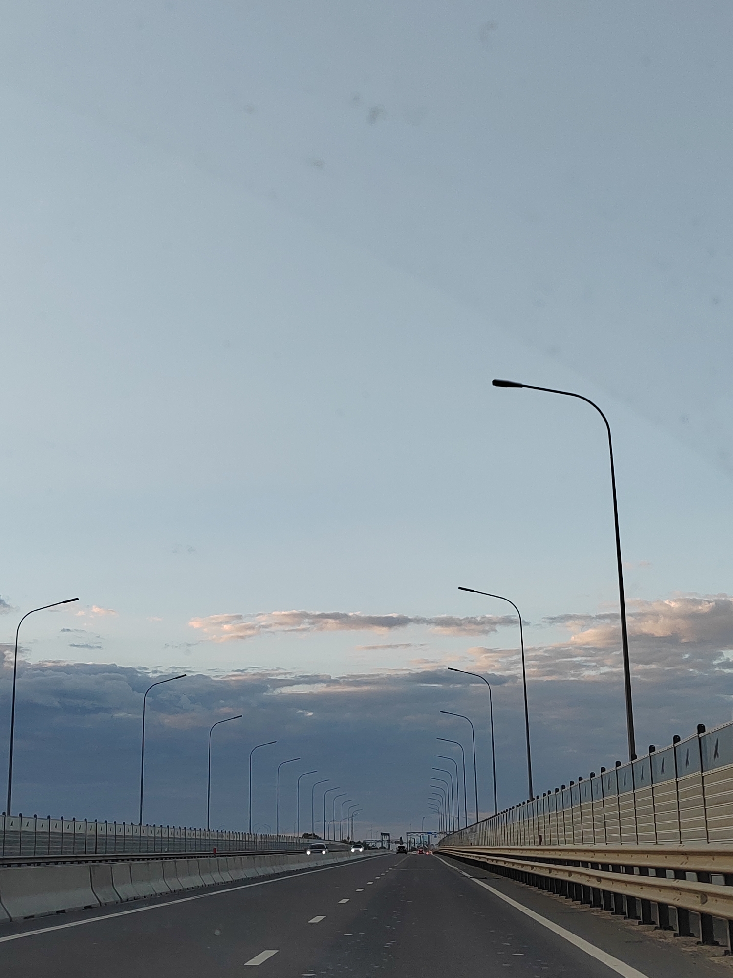 The image depicts a view from a vehicle traveling on a highway, likely a bridge, in the city of Brest, Belarus. The sky is a pale blue with scattered clouds tinged with pink and gray, suggesting it is either early morning or late evening. A series of tall, black streetlights line the right side of the highway, disappearing into the distance. The highway itself is asphalt with white lane markings. On the right, there is a concrete barrier with a metal railing above it, followed by a sound barrier. On the left side of the highway, there is a similar concrete barrier. Two vehicles are visible in the distance on the highway, their headlights on. The overall scene is devoid of any people, and the focus is on the infrastructure of the highway and the sky.