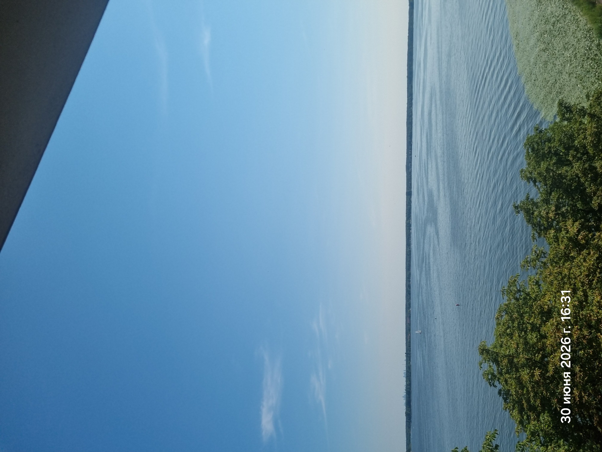 The image shows a wide expanse of a calm, blue lake under a clear blue sky. A thin line of trees and distant buildings marks the far shore. In the foreground, lush green trees partially obscure the lower portion of the scene. A timestamp in the bottom left corner reads "30 июня 2026 г. 16:31", which translates to "June 30, 2026, 16:31" in Russian. This suggests the image was taken in Zegrze, Poland, given the Russian text likely indicates a localized timestamp. The water surface is disturbed by gentle ripples, and a small sailboat is visible in the distance. There are no people or animals clearly identifiable in the image. The overall impression is one of a tranquil, sunny afternoon by the water.