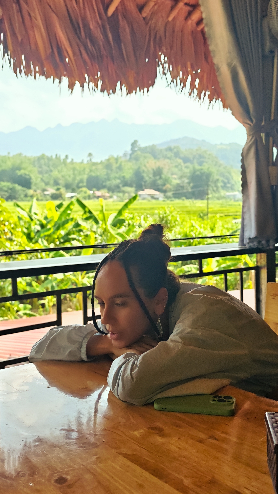 A woman rests her head on her hands at a polished wooden table. She is wearing a light-colored shirt and her hair is in a bun with braids framing her face. A green phone rests on the table beside her. The woman is looking down at the table. Behind her, a black metal railing separates the dining area from a green landscape of rice paddies and trees. In the distance, mountains are visible under a clear sky. A thatched roof hangs above the table. The location appears to be in Mậu Duệ Commune, Vietnam, based on the landscape and architectural style.